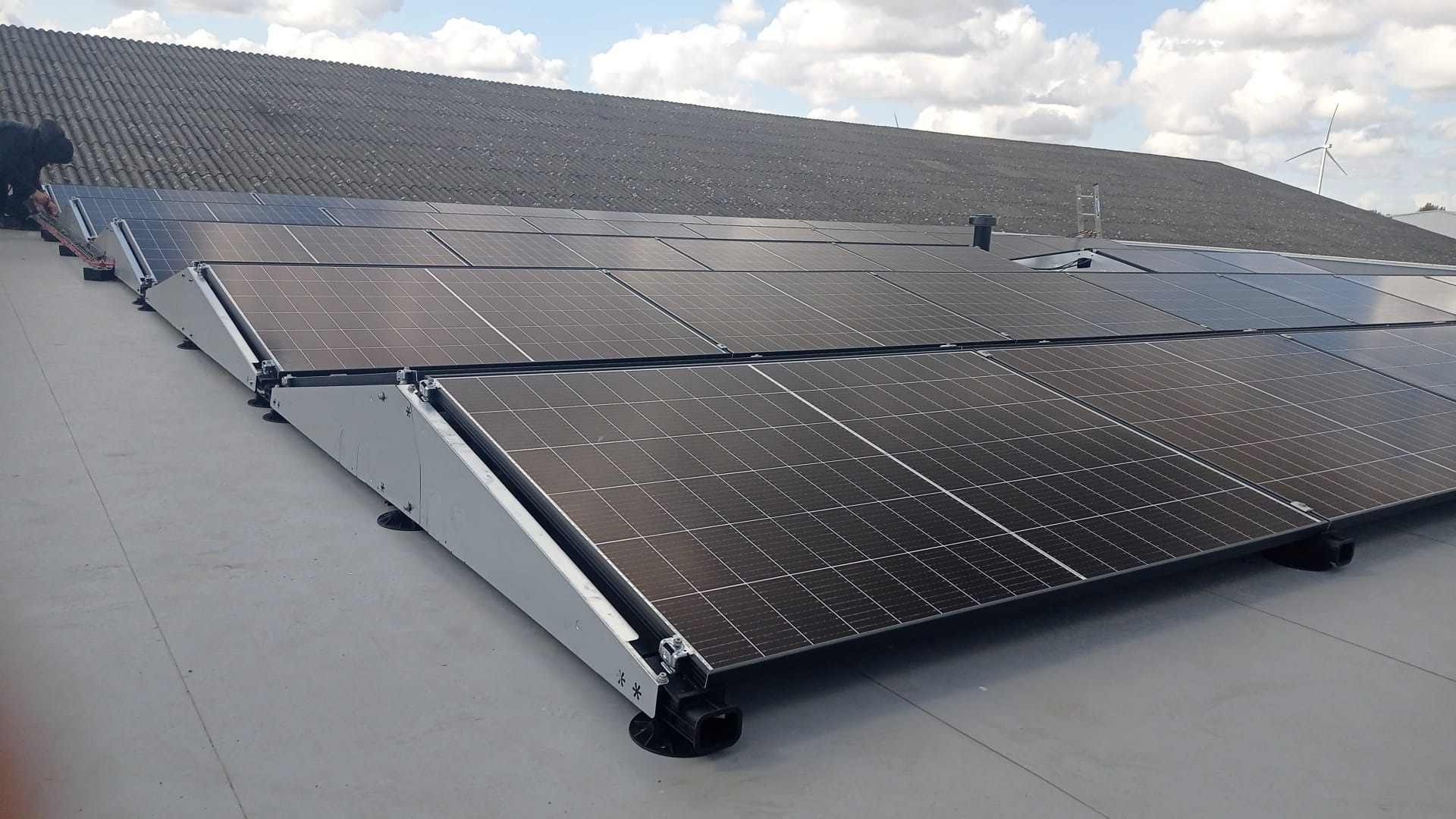 Montaż paneli fotowoltaicznych na płaskim dachu z widocznymi systemami mocowań i pracownikiem w lewym górnym rogu, w tle budynek z szarym dachem i turbina wiatrowa.