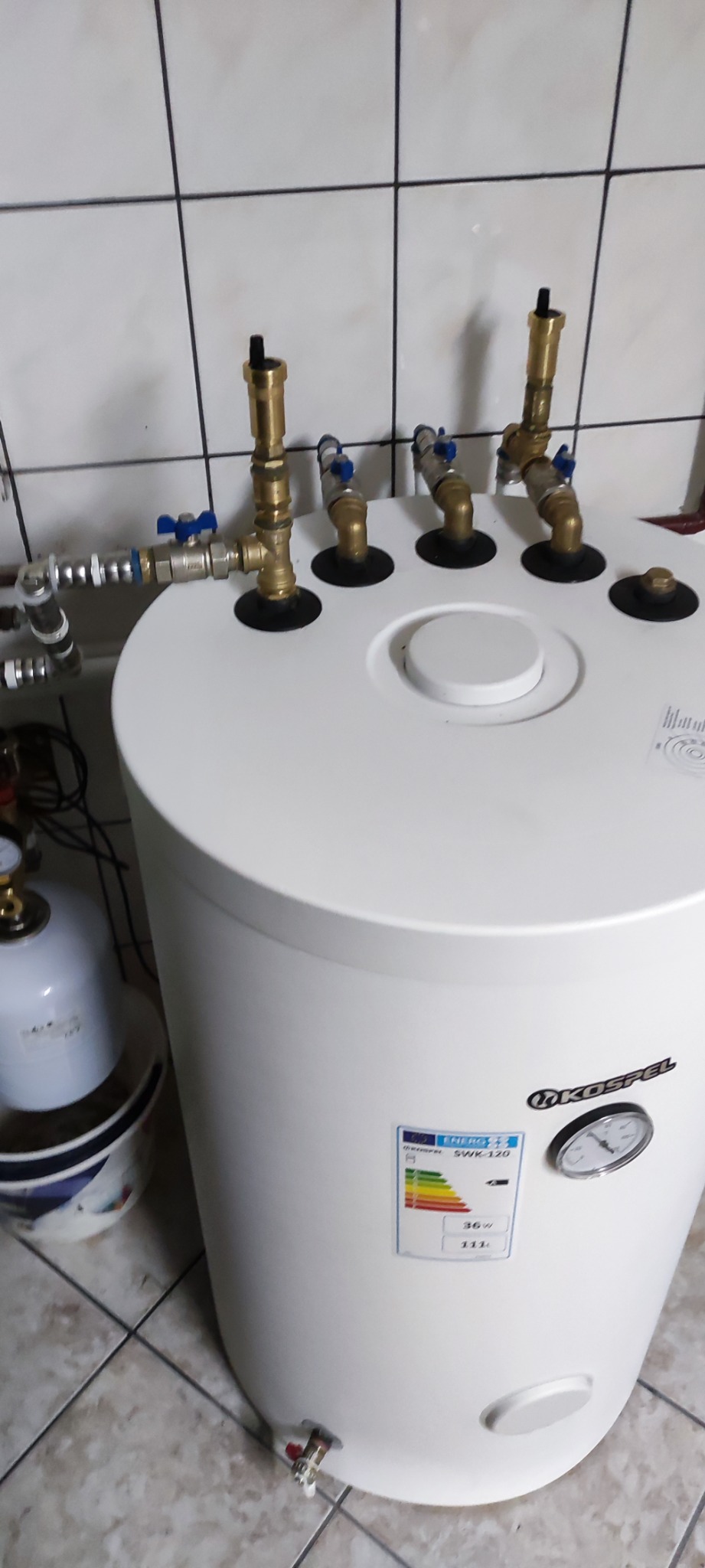 Bojler elektryczny Kospel SWK-120 z podłączonymi zaworami i rurami miedzianymi, widoczna etykieta energetyczna i manometr, na tle białych płytek.