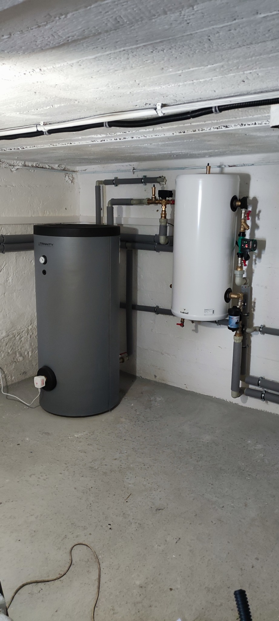 Szary zbiornik buforowy Trinity i biały zasobnik ciepłej wody Galmet w piwnicy z rurami instalacyjnymi pod sufitem.