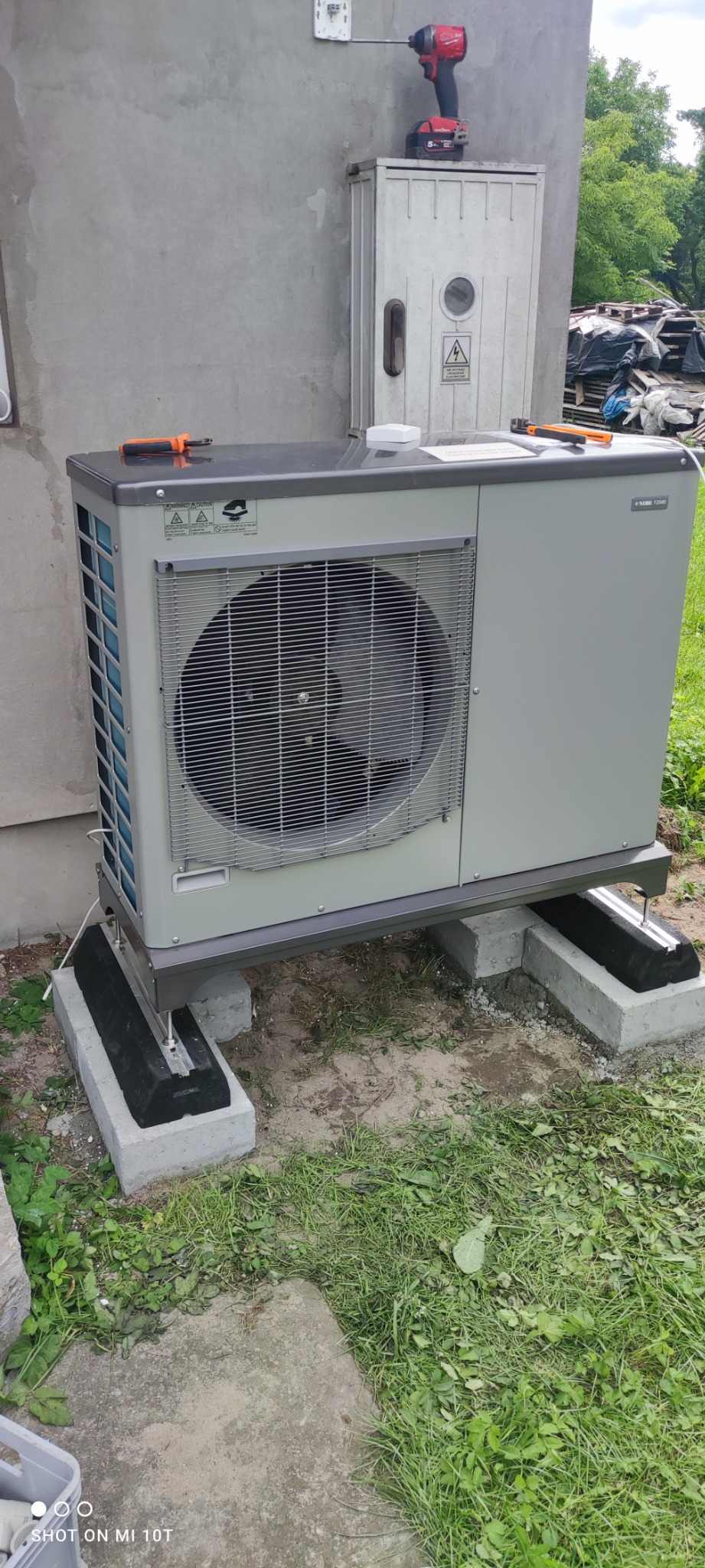 Zewnętrzna jednostka pompy ciepła zamontowana na betonowych blokach, z widoczną skrzynką elektryczną i wkrętarką na górze, na tle trawy i ściany budynku.