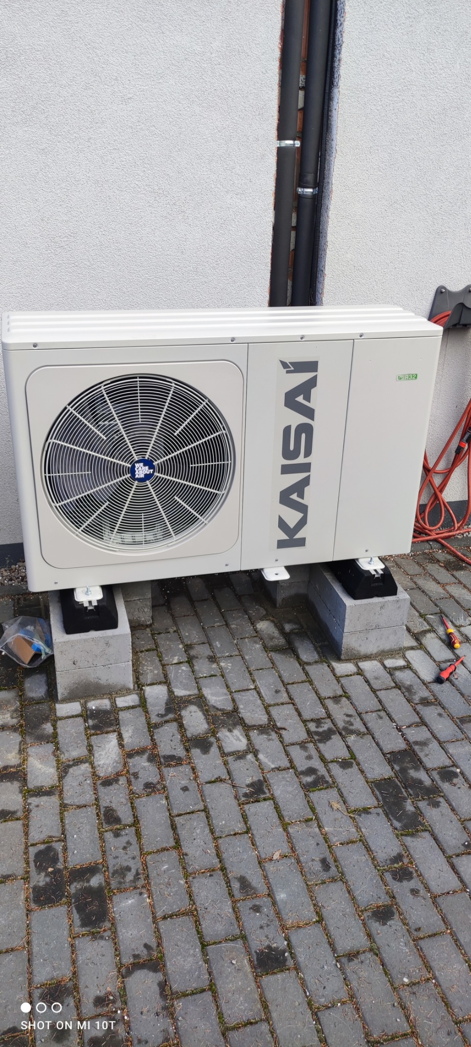 Biała jednostka zewnętrzna pompy ciepła marki Kaisai, zamontowana na betonowych bloczkach na szarej kostce brukowej, widoczne narzędzia i przewody w tle.
