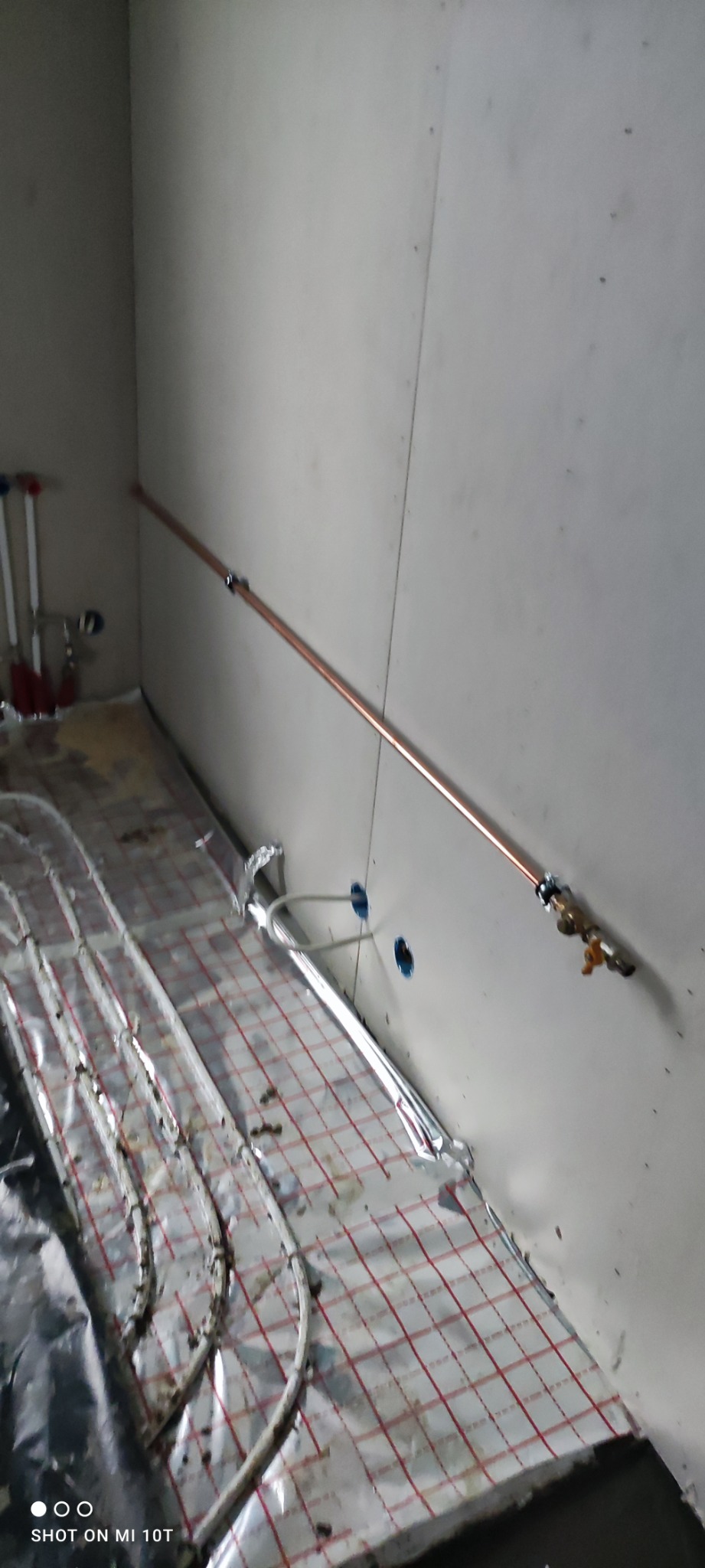 Instalacja ogrzewania podłogowego z rurami pokrytymi folią aluminiową na podkładzie z siatką, widoczne rury miedziane na ścianie z zaworami.