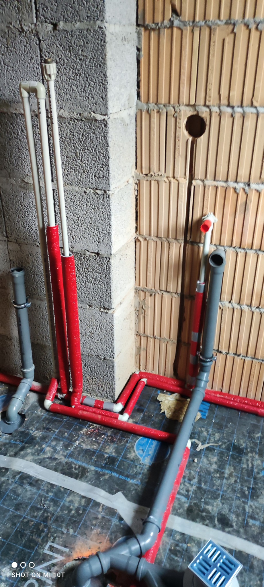 Wewnętrzna instalacja wodno-kanalizacyjna w trakcie montażu: widoczne rury PCV w kolorze białym i czerwonym, szare rury kanalizacyjne oraz kratka ściekowa na podłodze z płytek. Ściany z pustaków...