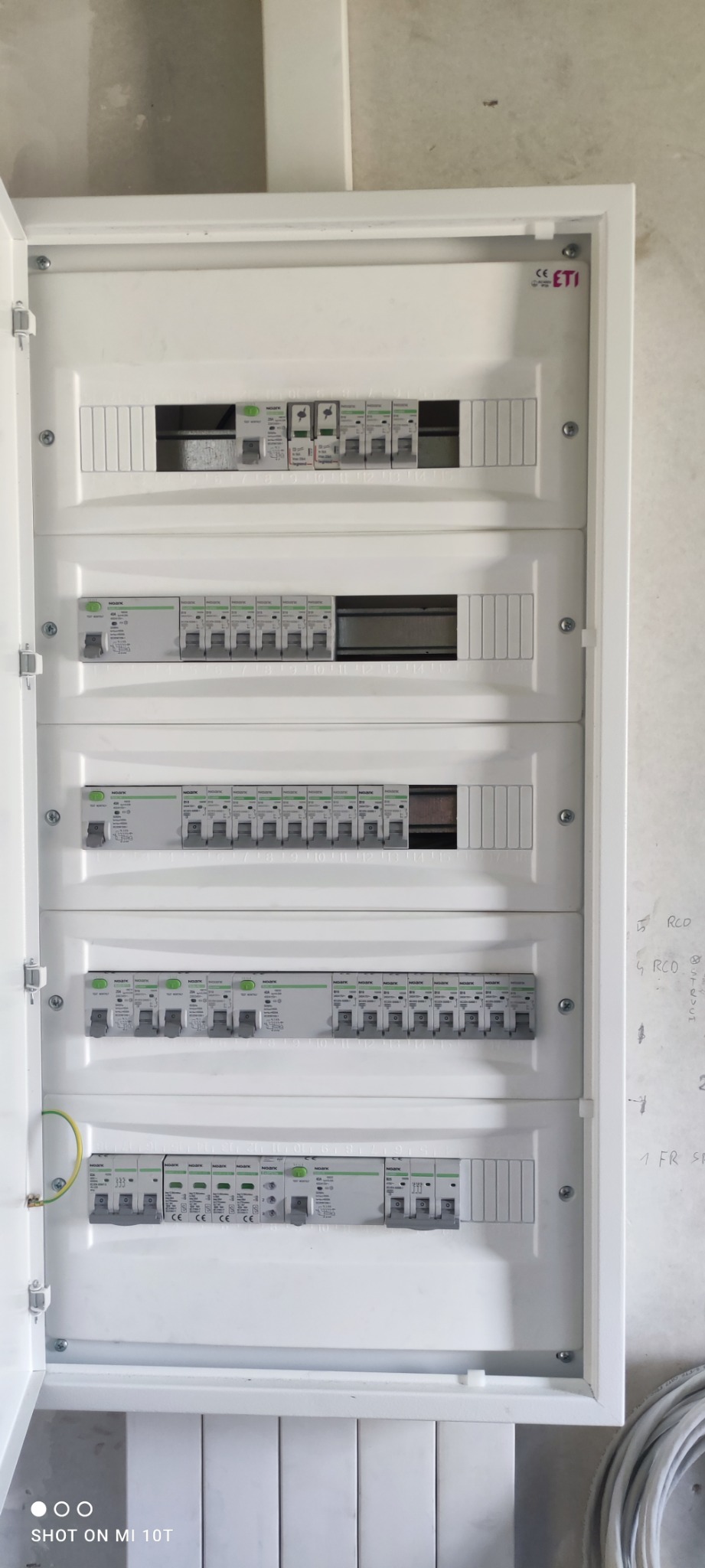 Otwarta, biała skrzynka elektryczna z zamontowanymi bezpiecznikami NOARK i wyłącznikami różnicowoprądowymi, widoczne przewody. Na ścianie obok notatki ołówkiem.