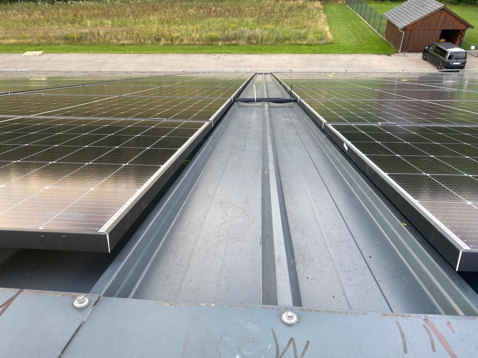 Panele fotowoltaiczne zamontowane na blaszanym dachu z widoczną przestrzenią między rzędami paneli, w tle budynek gospodarczy i zaparkowany samochód.