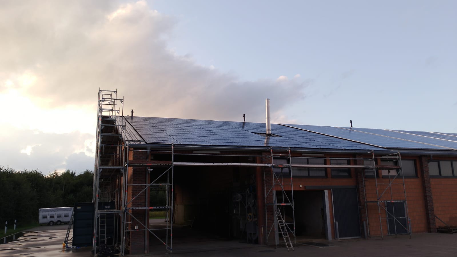 Montaż paneli fotowoltaicznych na dachu budynku z cegły, widoczne rusztowania i pochmurne niebo.