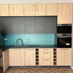 Nowoczesna kuchnia z drewnianymi frontami szafek górnych i dolnych, szarymi szafkami wiszącymi, turkusowymi płytkami na ścianie i podłogą w jodełkę.