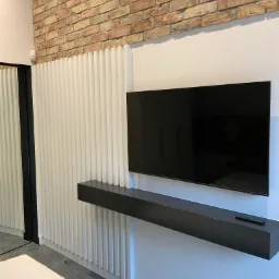 Nowoczesna aranżacja ściany z telewizorem: białe, pionowe panele dekoracyjne, ceglana ściana w tle, czarna półka z pilotem pod telewizorem.