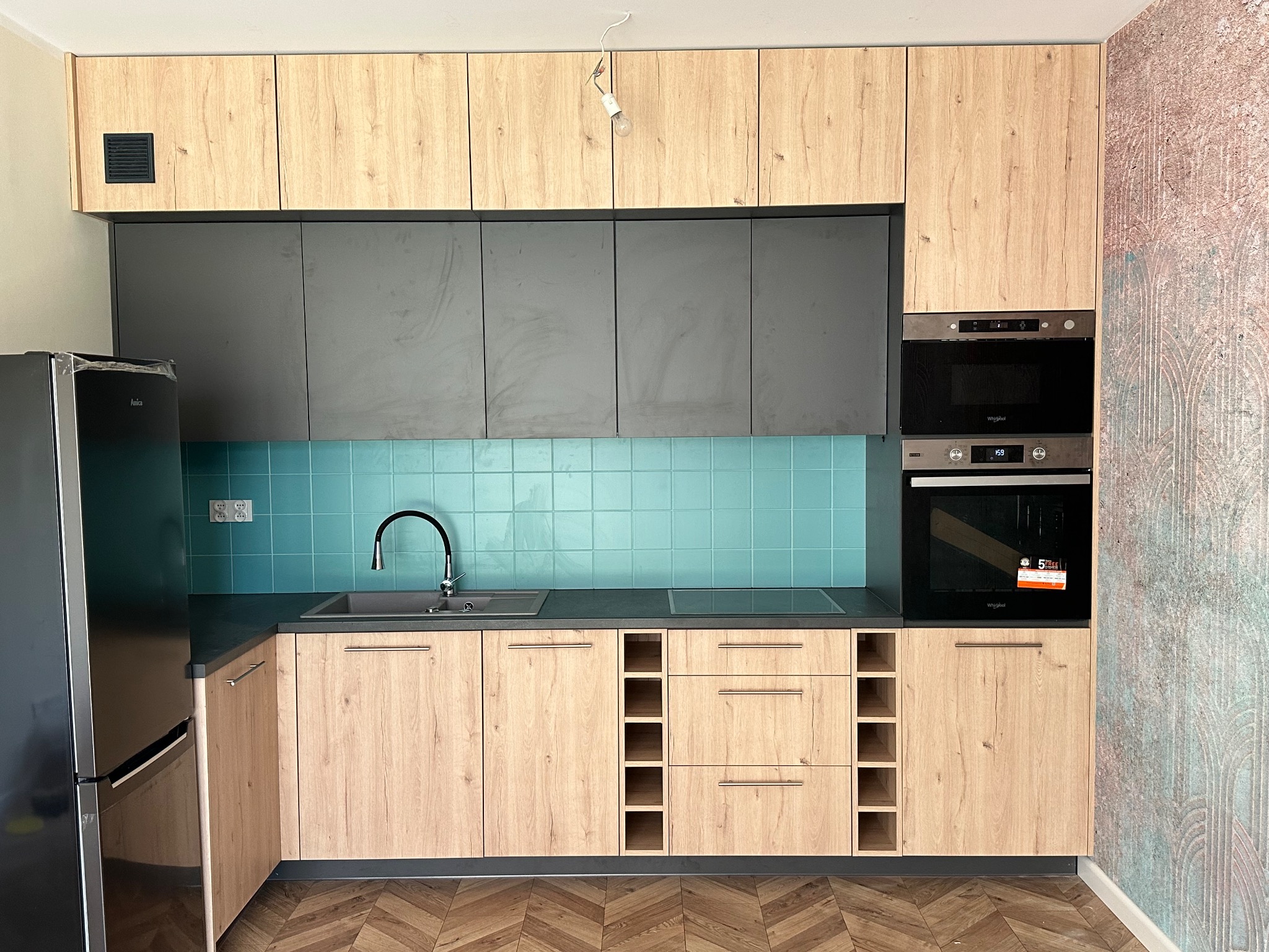 Nowoczesna kuchnia z drewnianymi frontami szafek górnych i dolnych, szarymi szafkami wiszącymi, turkusowymi płytkami na ścianie i podłogą w jodełkę.