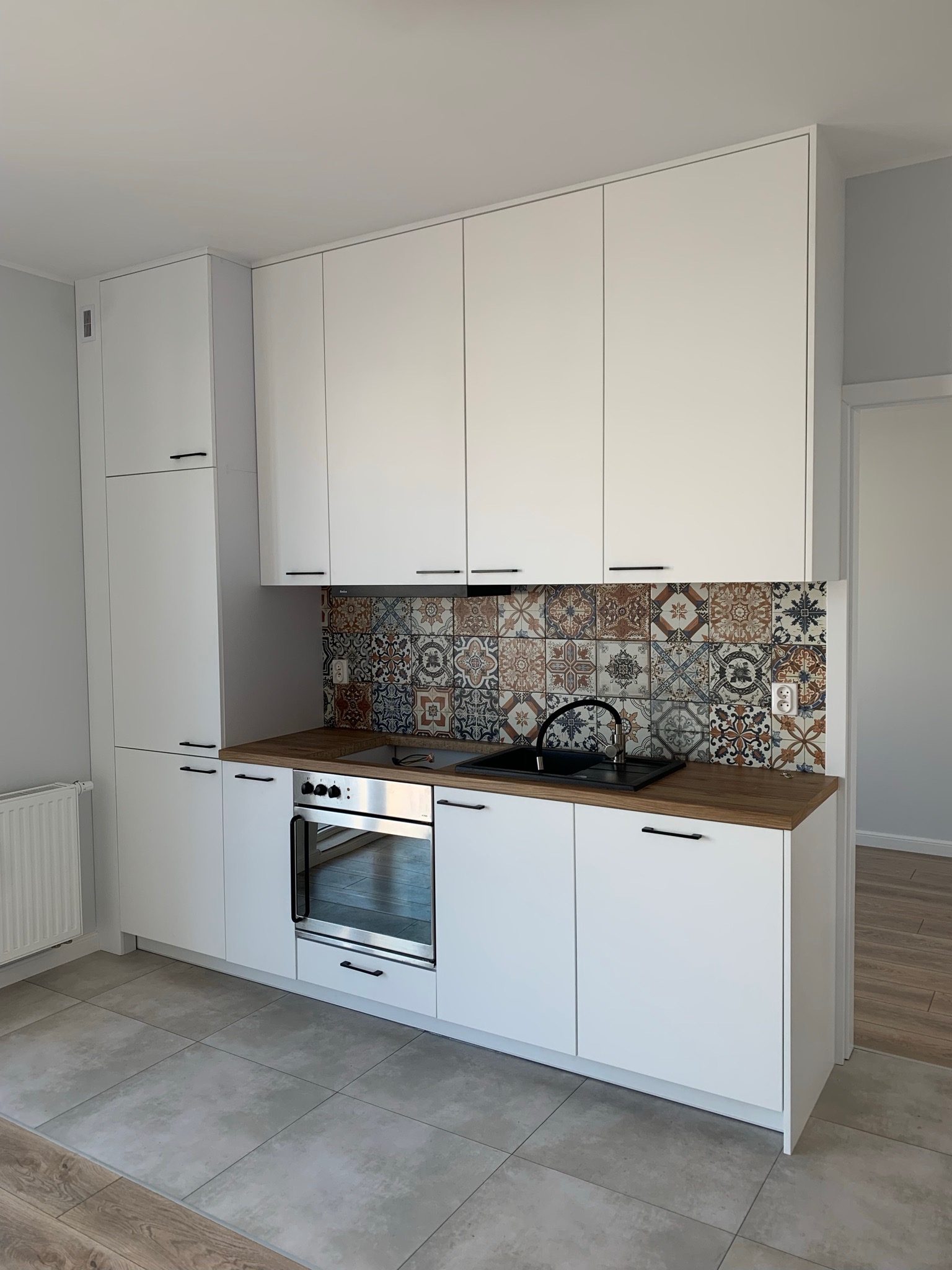 Nowoczesna kuchnia z białymi, matowymi frontami, drewnianym blatem i dekoracyjną mozaiką nad blatem, czarny zlew i bateria, piekarnik w zabudowie.