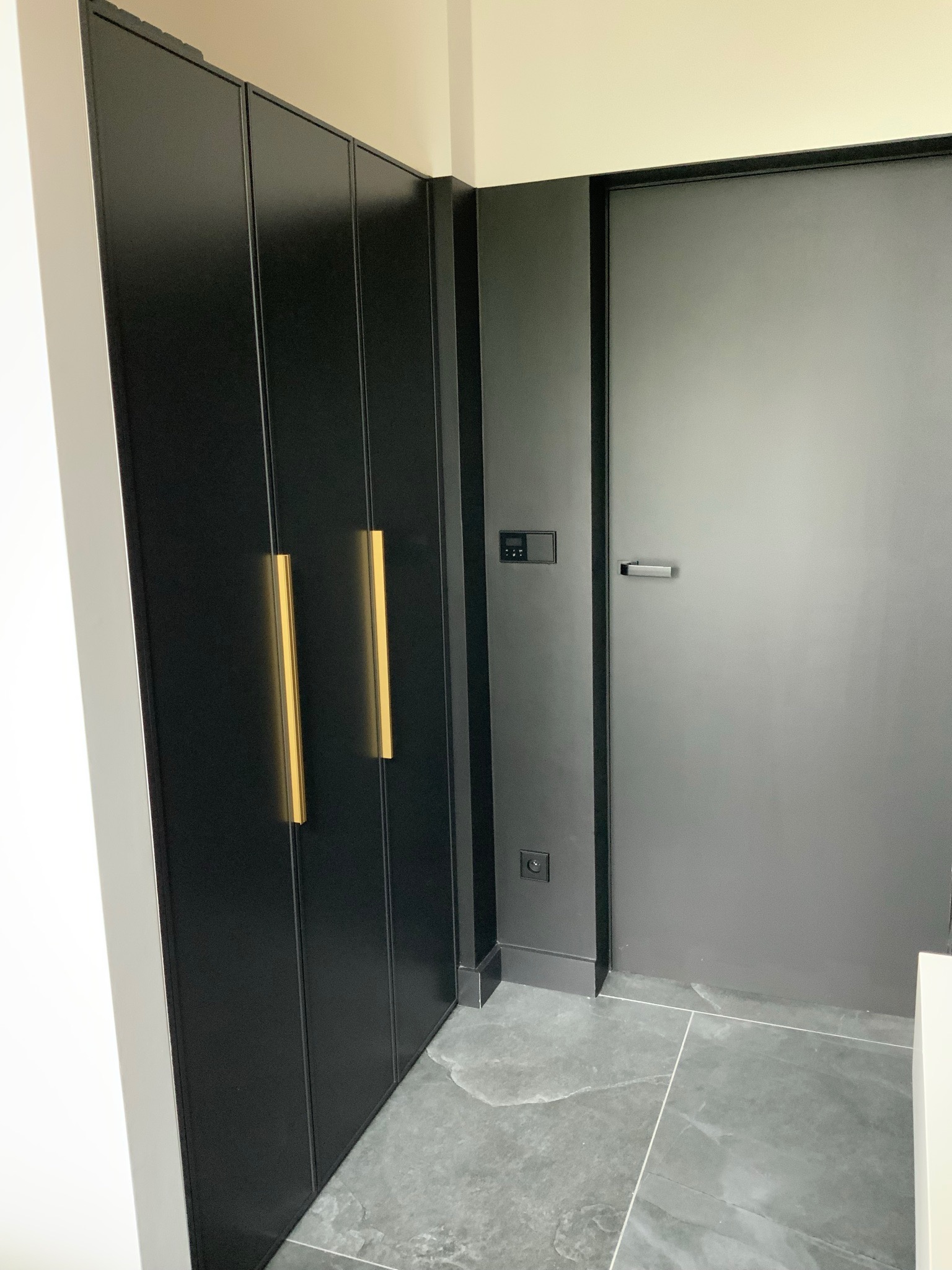 Czarna szafa na wymiar z pionowymi złotymi uchwytami, sąsiadująca z szarą ścianą i drzwiami na tle szarej marmurowej podłogi.