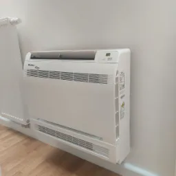 Biała klimatyzacja podłogowa marki Gree zamontowana przy ścianie obok grzejnika, widoczny wyświetlacz z temperaturą.