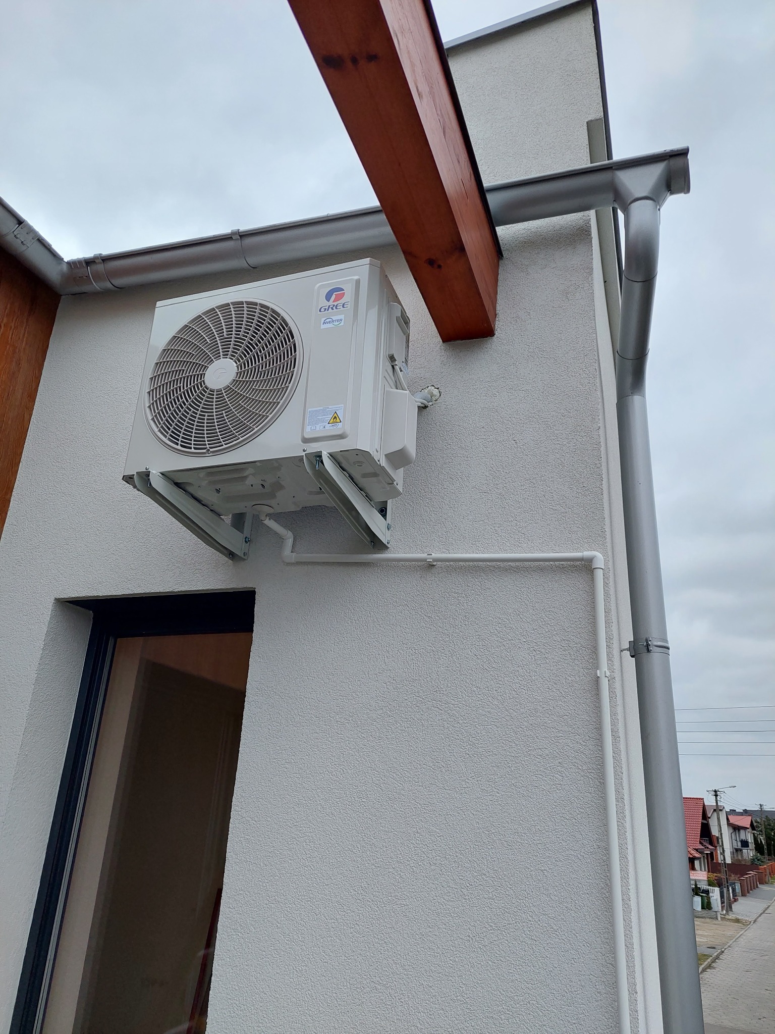 Zewnętrzna jednostka klimatyzacji marki Gree zamontowana na białej elewacji domu, widoczne rynny i drewniana konstrukcja dachu.