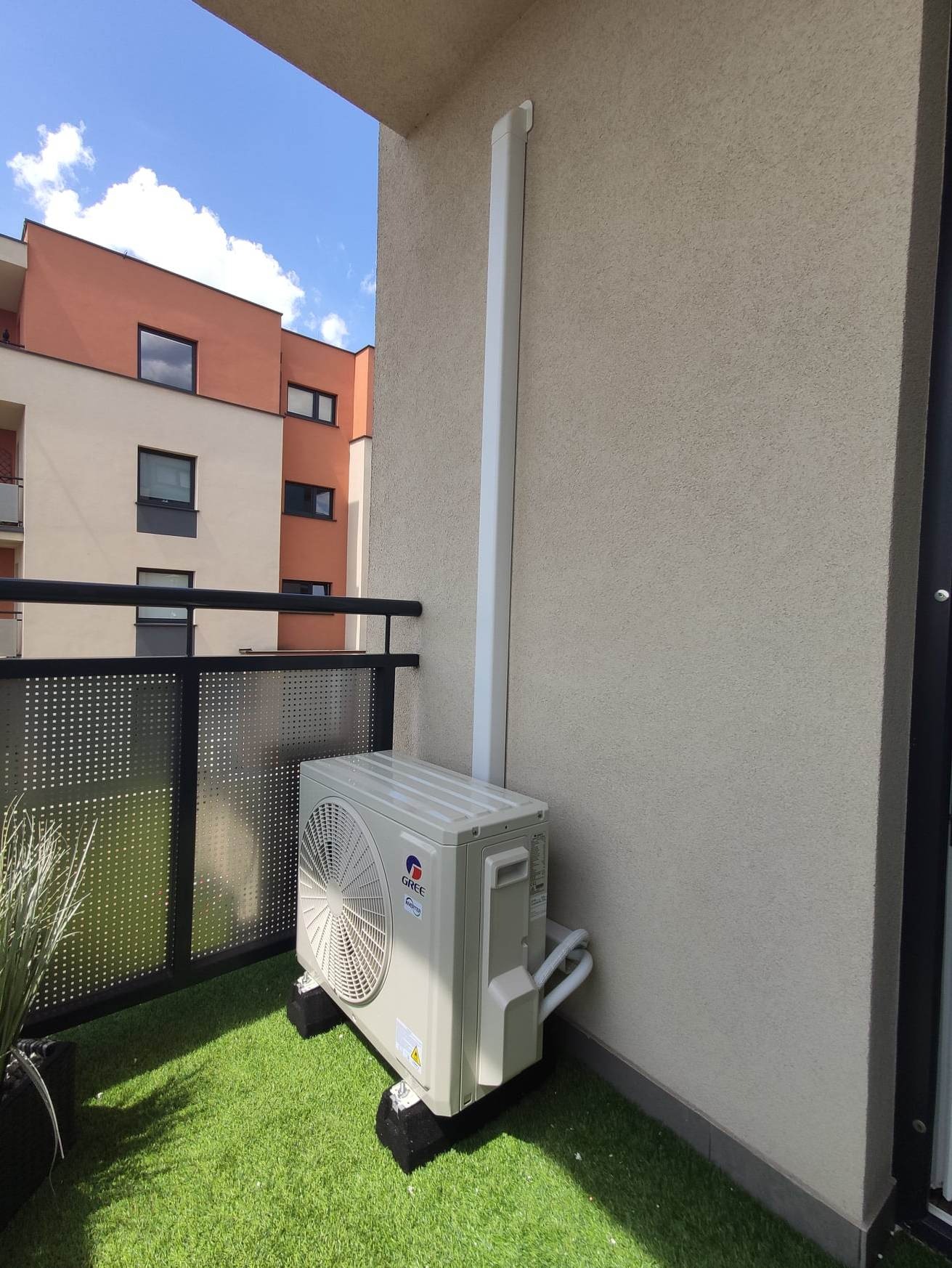 Jednostka zewnętrzna klimatyzatora marki Gree zamontowana na balkonie z imitacją trawy, widoczna rura odprowadzająca skropliny, w tle budynek mieszkalny.