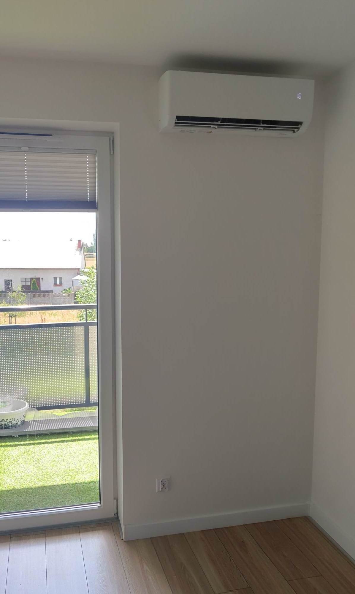 Biały klimatyzator ścienny zamontowany w pokoju z oknem balkonowym i drewnianą podłogą; widoczny wyświetlacz temperatury.