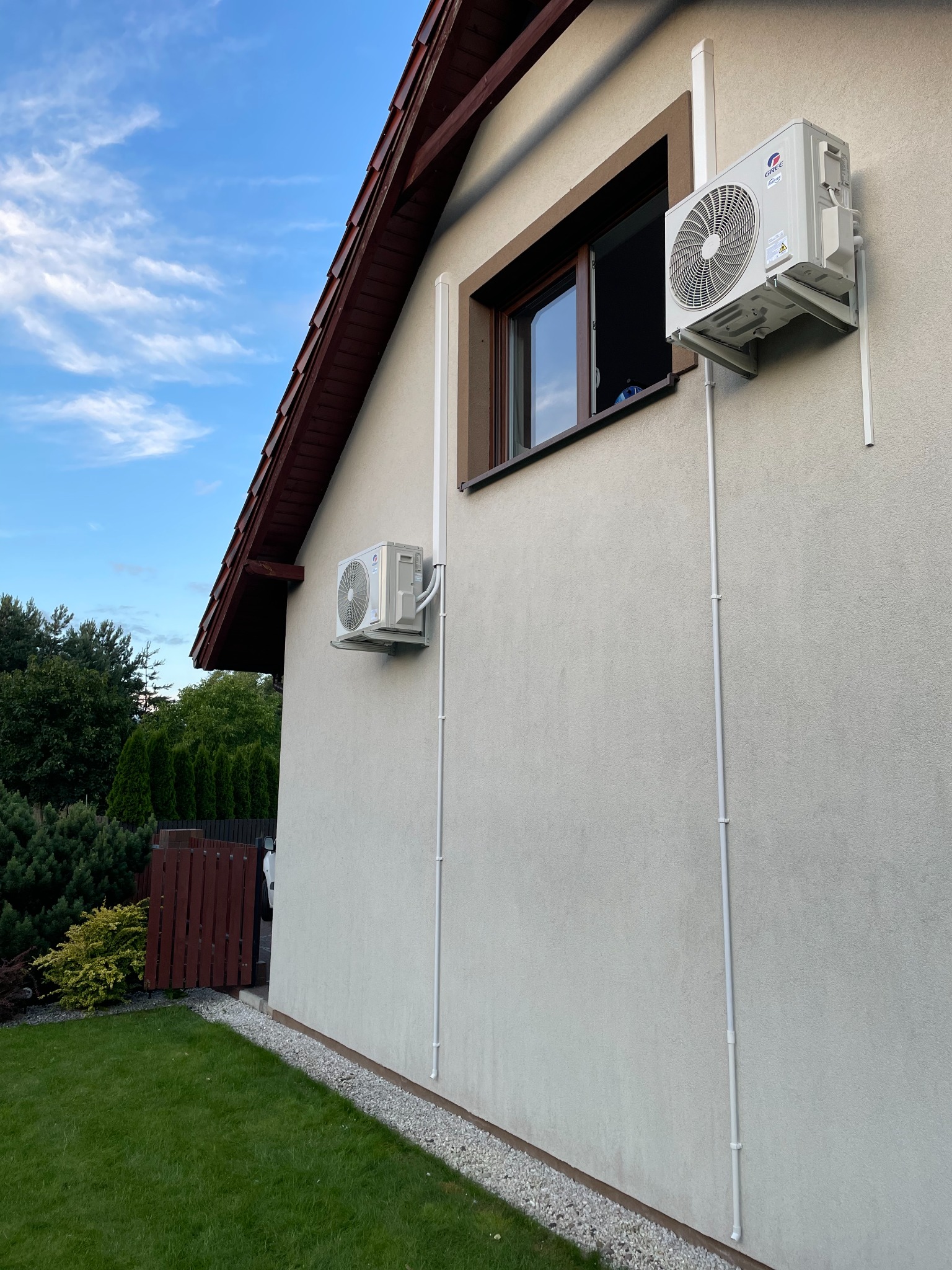 Dwa klimatyzatory marki Gree zamontowane na elewacji domu jednorodzinnego, widoczne rury odprowadzające skropliny i przewody elektryczne, tło: błękitne niebo z chmurami i zieleń drzew.