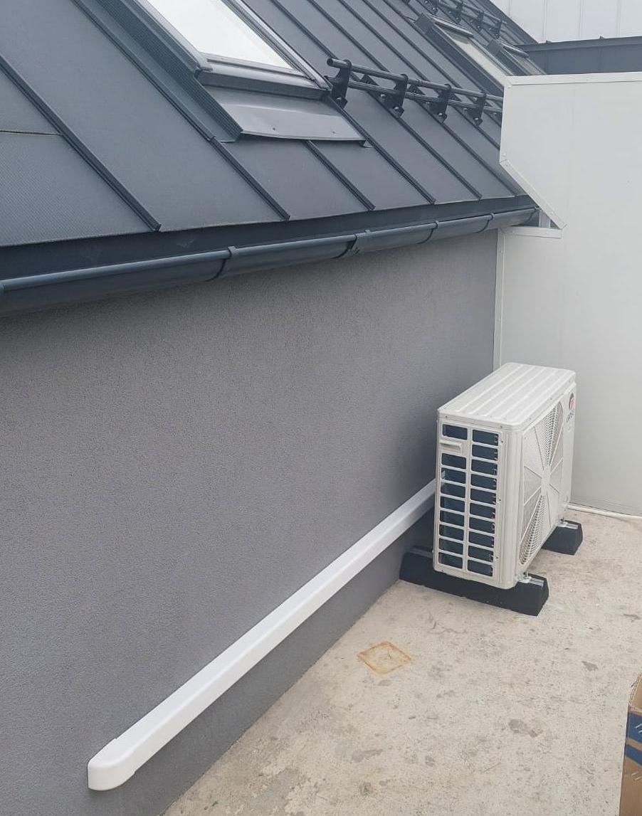 Zewnętrzna jednostka klimatyzacji zamontowana na betonowej podstawie przy szarej ścianie budynku z widoczną rynną i oknem dachowym.