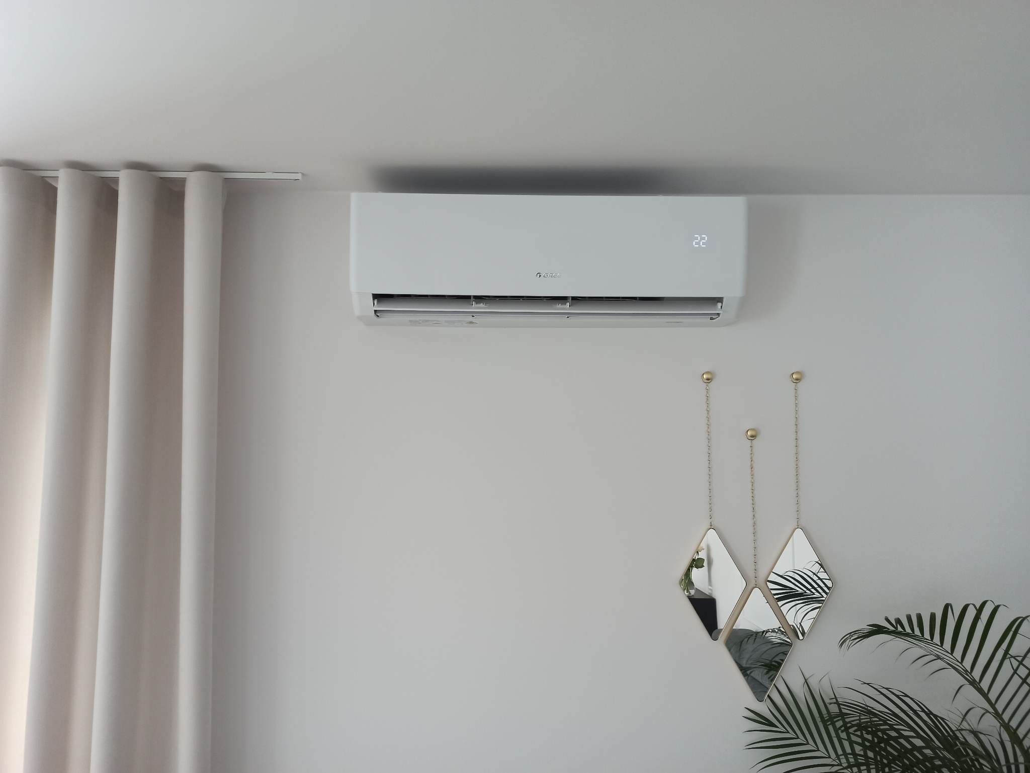 Biały klimatyzator ścienny marki Gree z wyświetlaczem temperatury 22 stopnie Celsjusza w minimalistycznym wnętrzu z zasłonami i dekoracyjnymi lustrami.