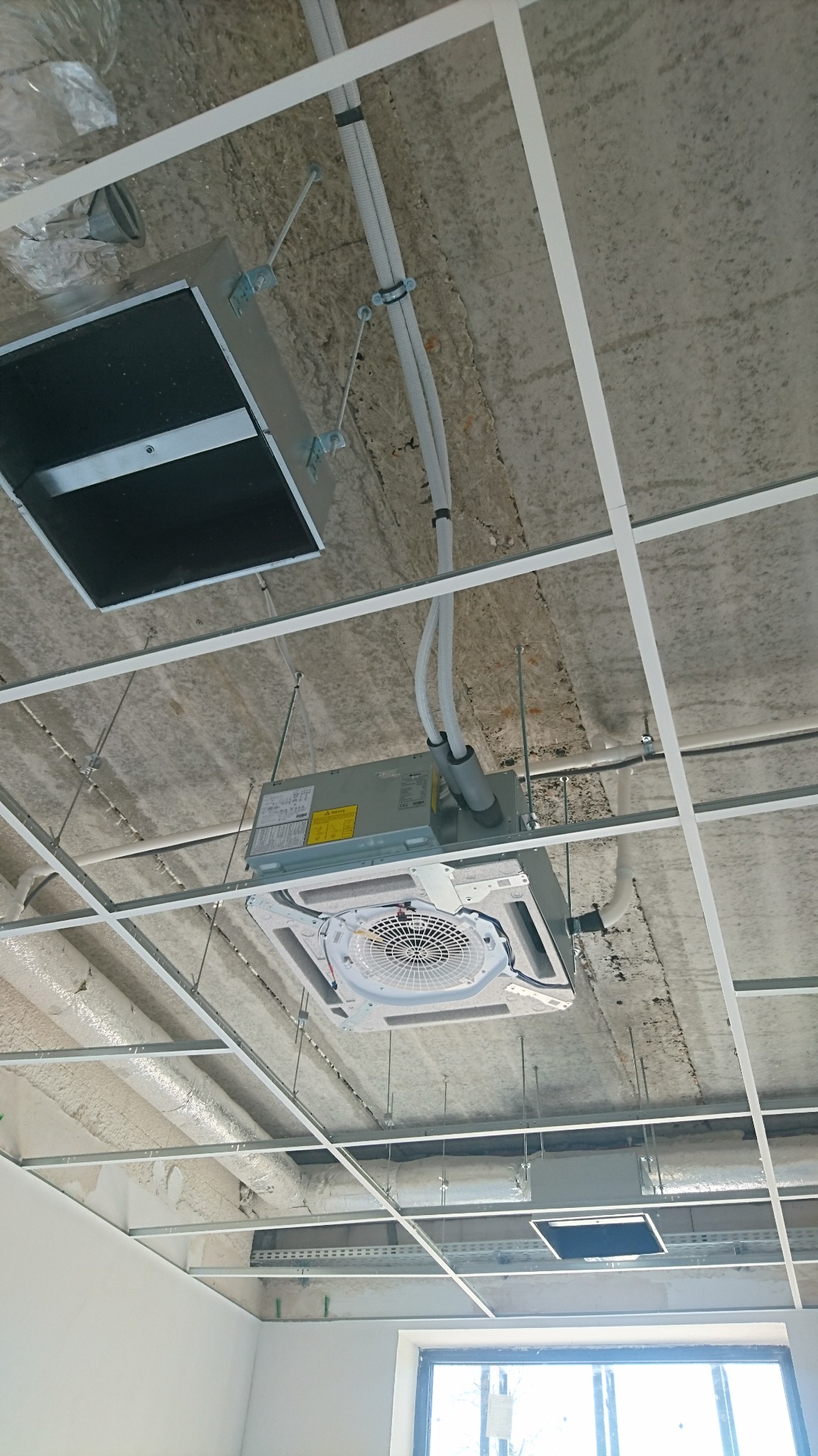 Montaż sufitowej jednostki klimatyzacyjnej w pomieszczeniu z surowym betonowym stropem i widoczną konstrukcją sufitu podwieszanego.
