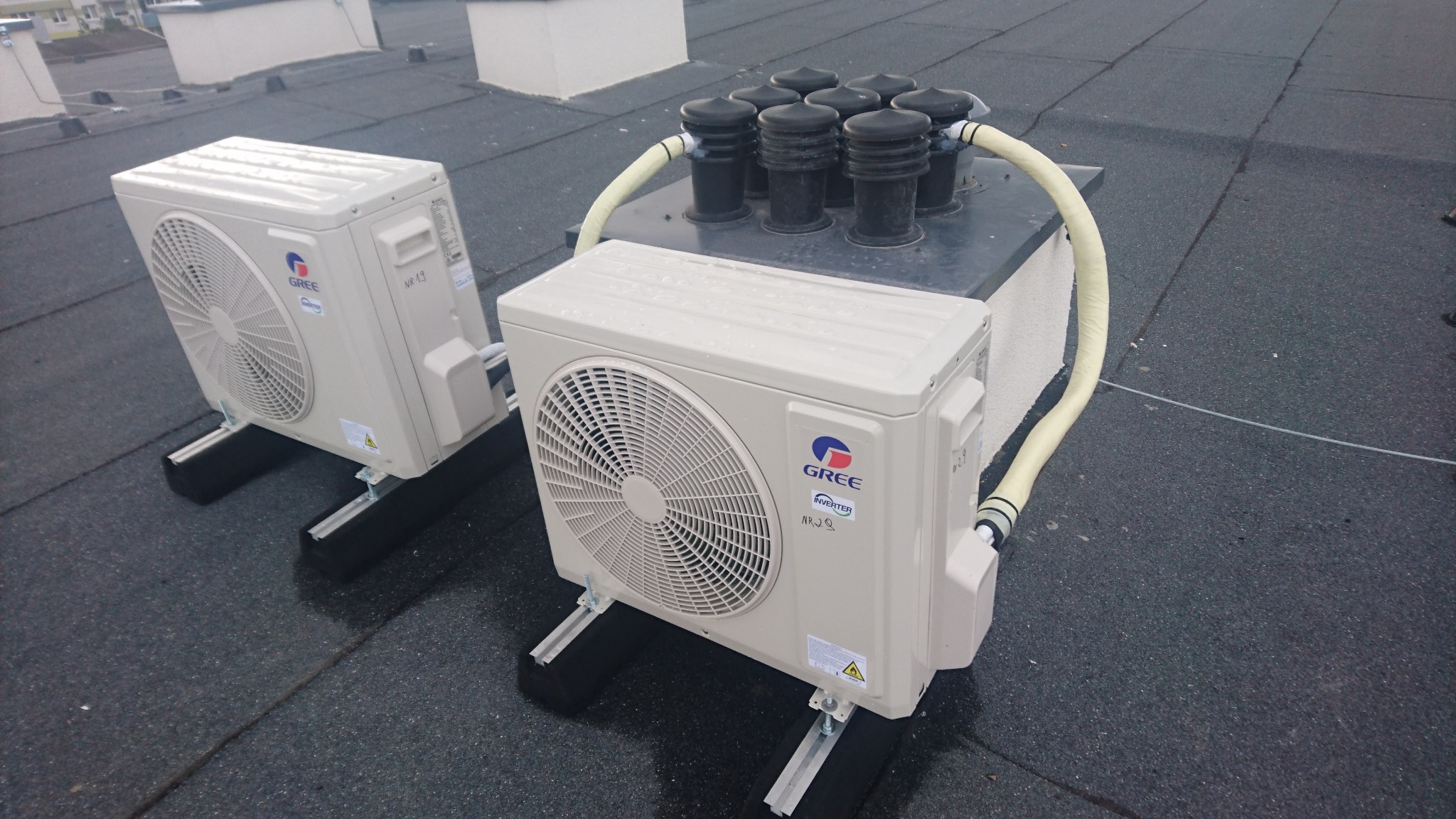 Dwie jednostki zewnętrzne klimatyzacji marki Gree na płaskim, bitumicznym dachu, z widocznymi przewodami i systemem odprowadzania skroplin.