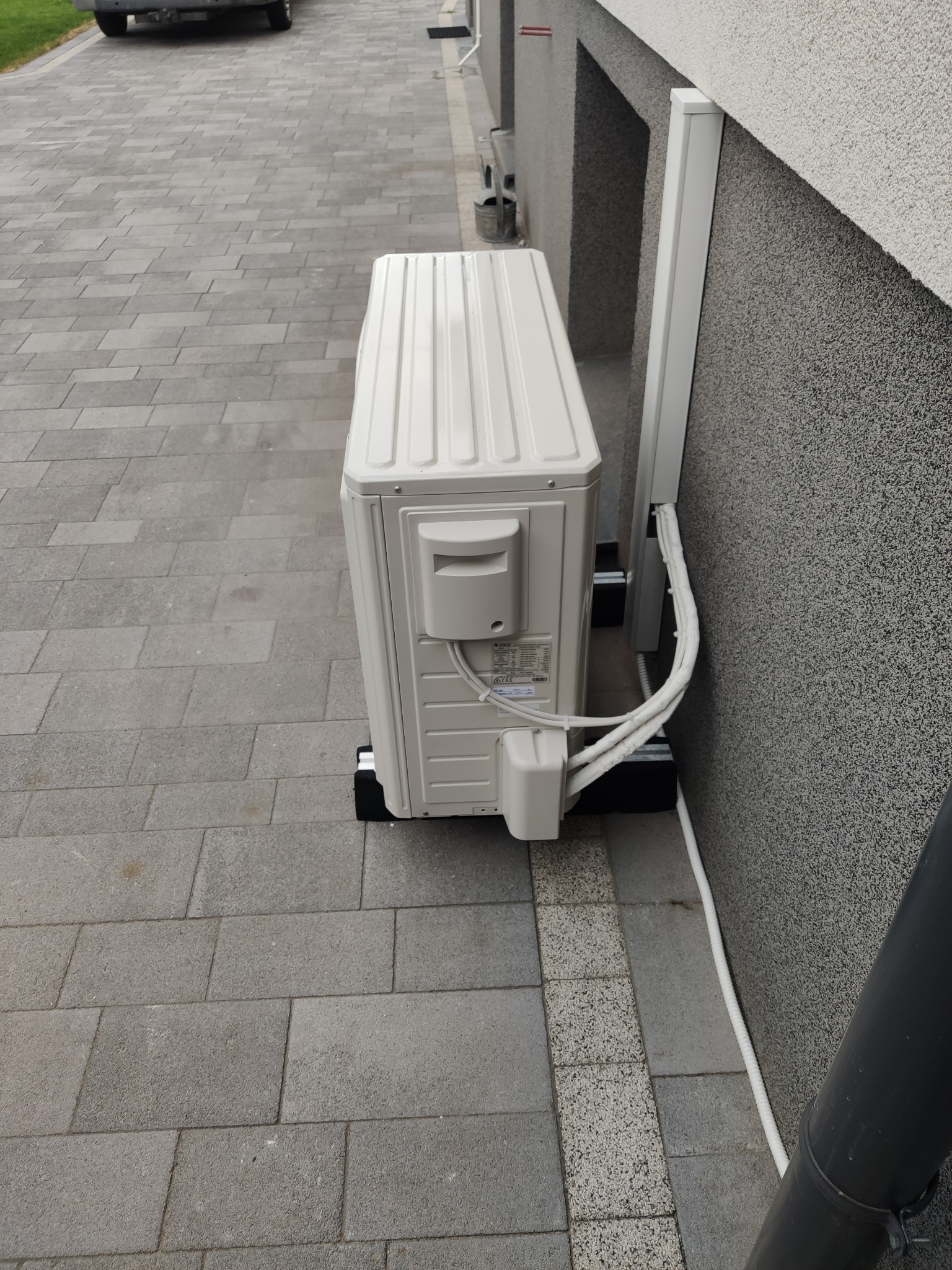 Zewnętrzna jednostka klimatyzacji zamontowana przy ścianie budynku z widocznymi przewodami w osłonie i kostką brukową w tle.