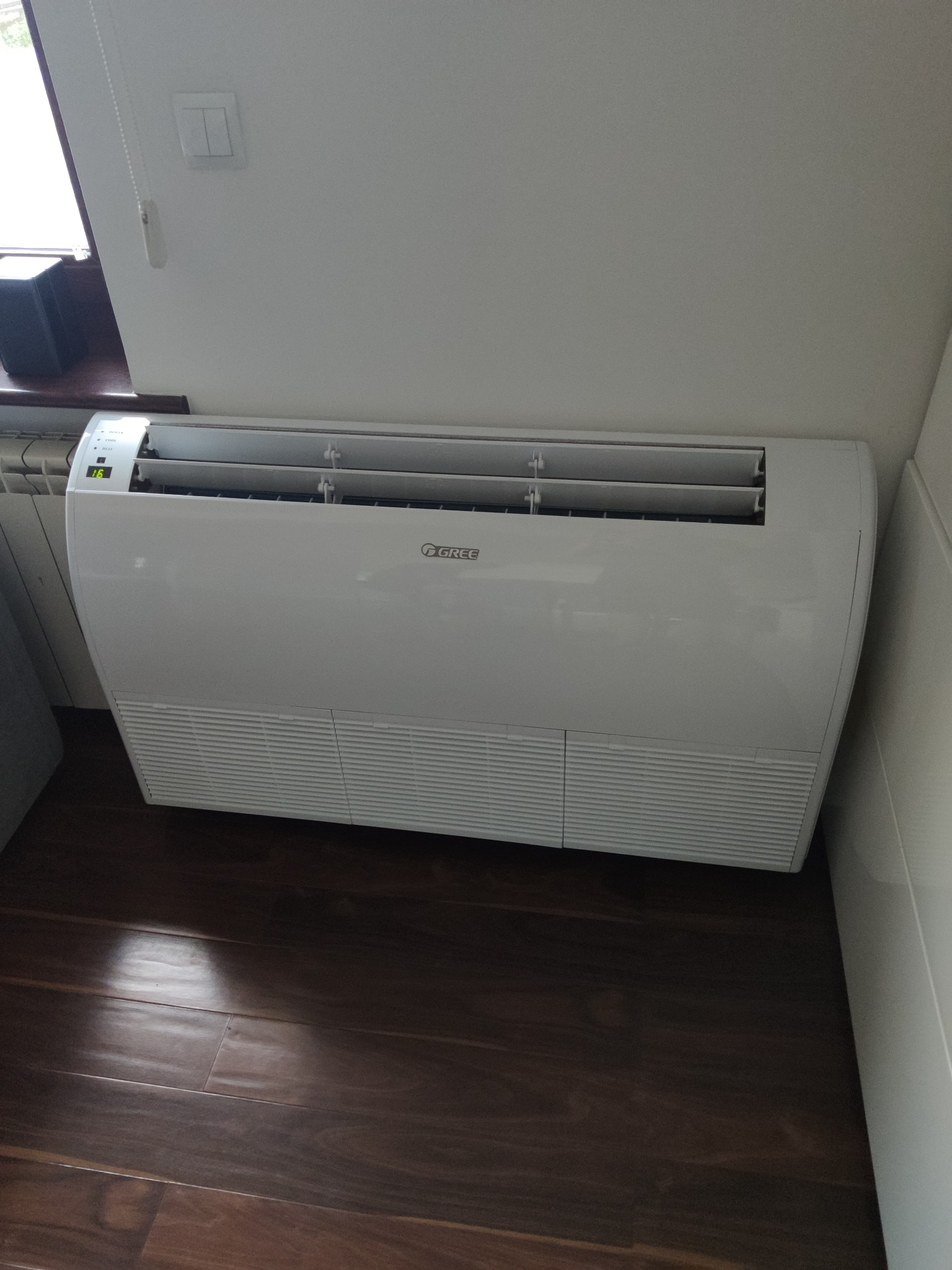 Biały klimatyzator przypodłogowy marki Gree zamontowany w pomieszczeniu z brązową podłogą, widoczny wyświetlacz temperatury ustawiony na 16 stopni.