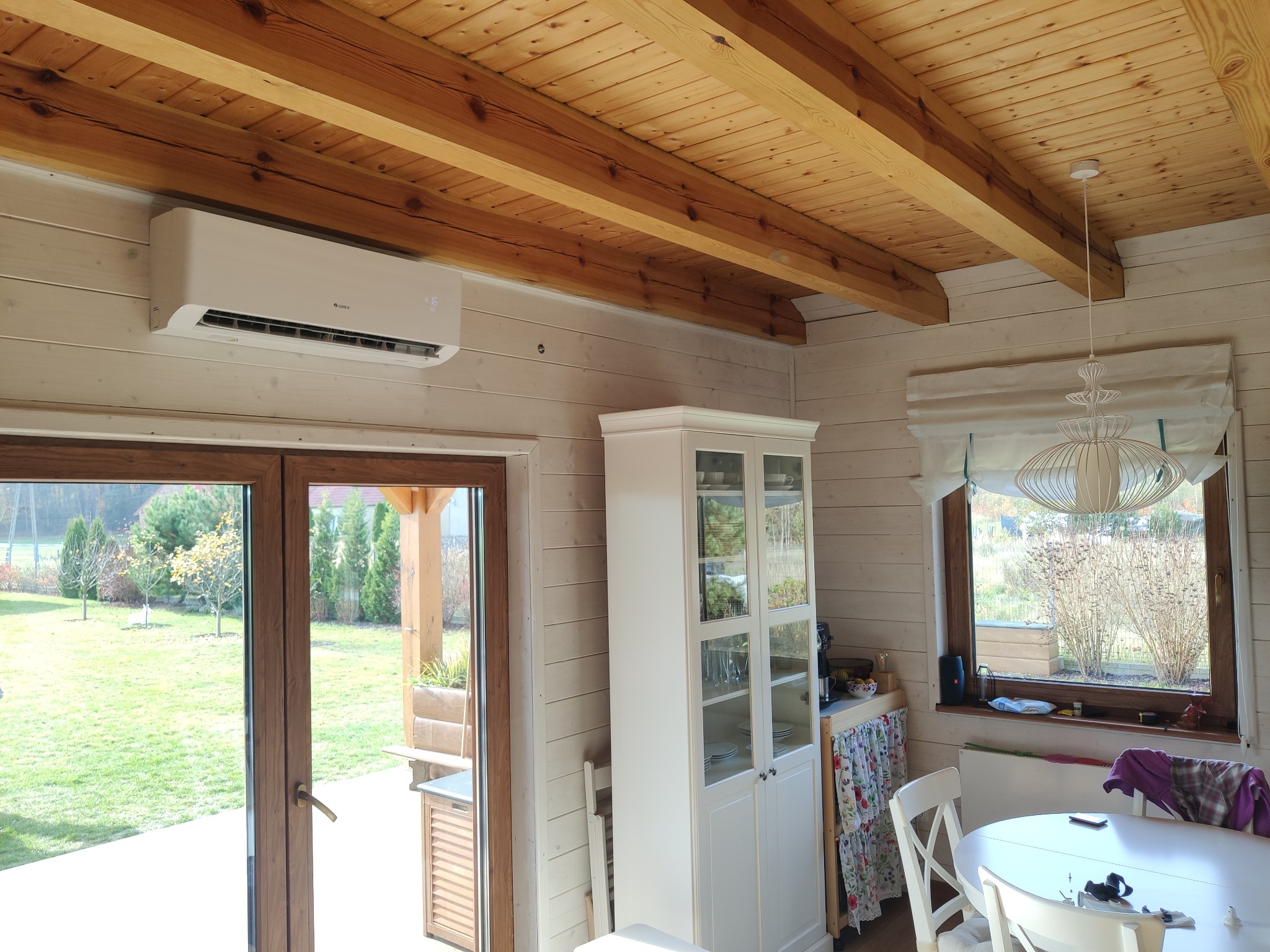 Biała jednostka klimatyzacyjna marki Gree zamontowana na ścianie w jasnym, drewnianym pokoju z widokiem na ogród przez otwarte drzwi balkonowe.
