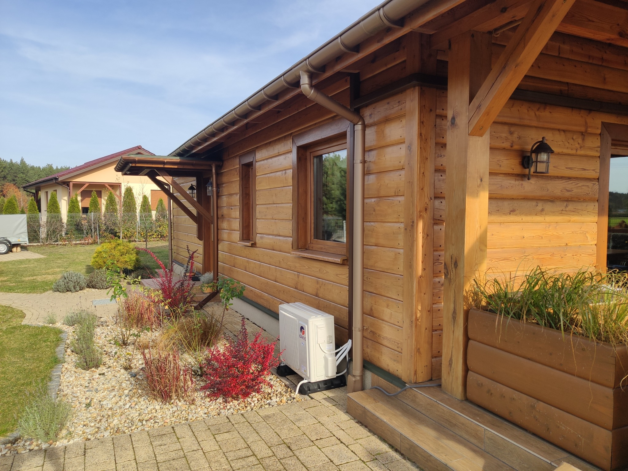 Zewnętrzna jednostka klimatyzacji marki Gree zamontowana przy drewnianym domu z brązowymi oknami, widoczne rośliny ozdobne i kostka brukowa.