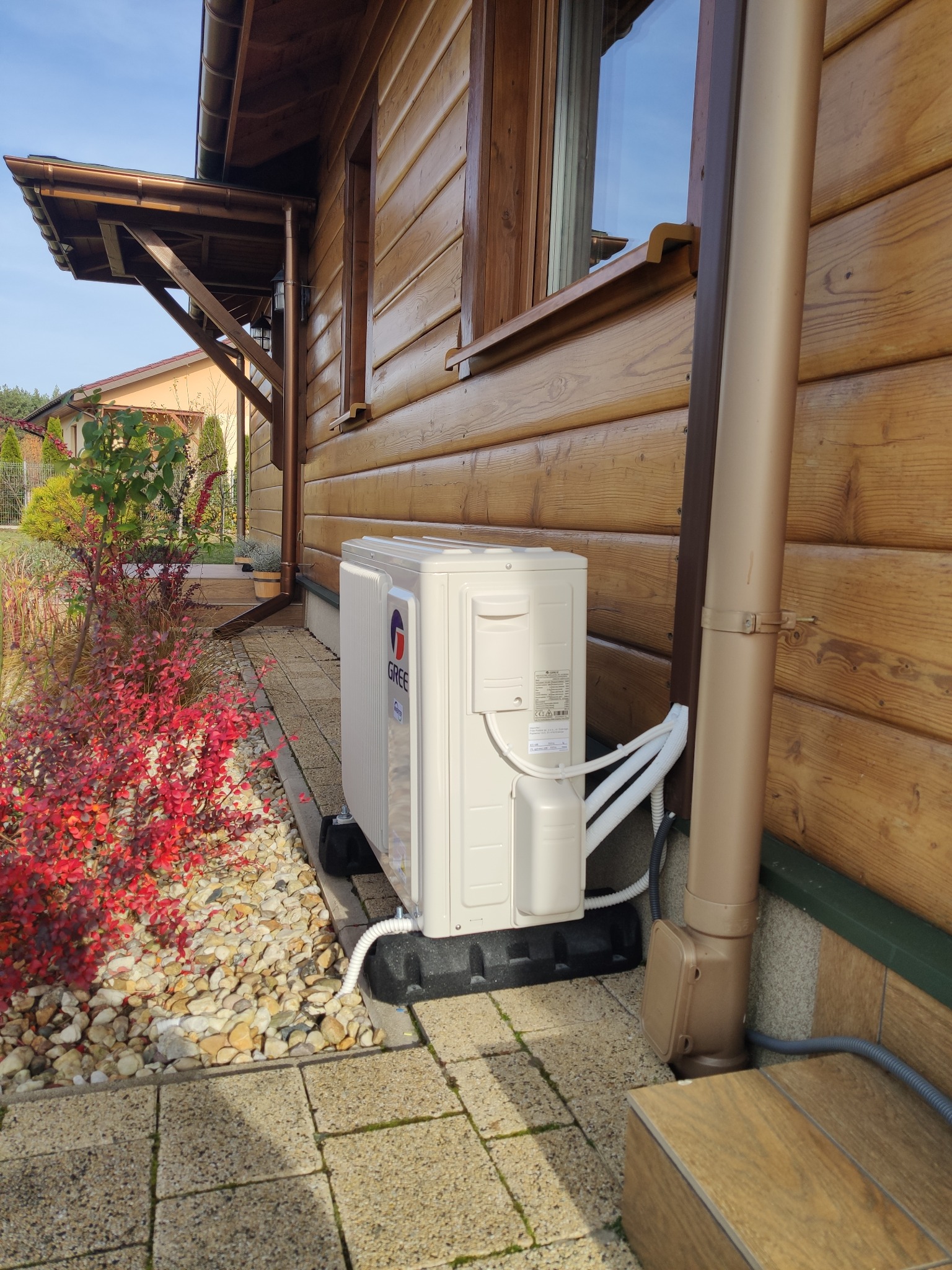 Zewnętrzna jednostka klimatyzacji marki Gree zamontowana przy drewnianym domu, widoczne przewody, rynna odprowadzająca wodę i rabata z czerwonymi krzewami.