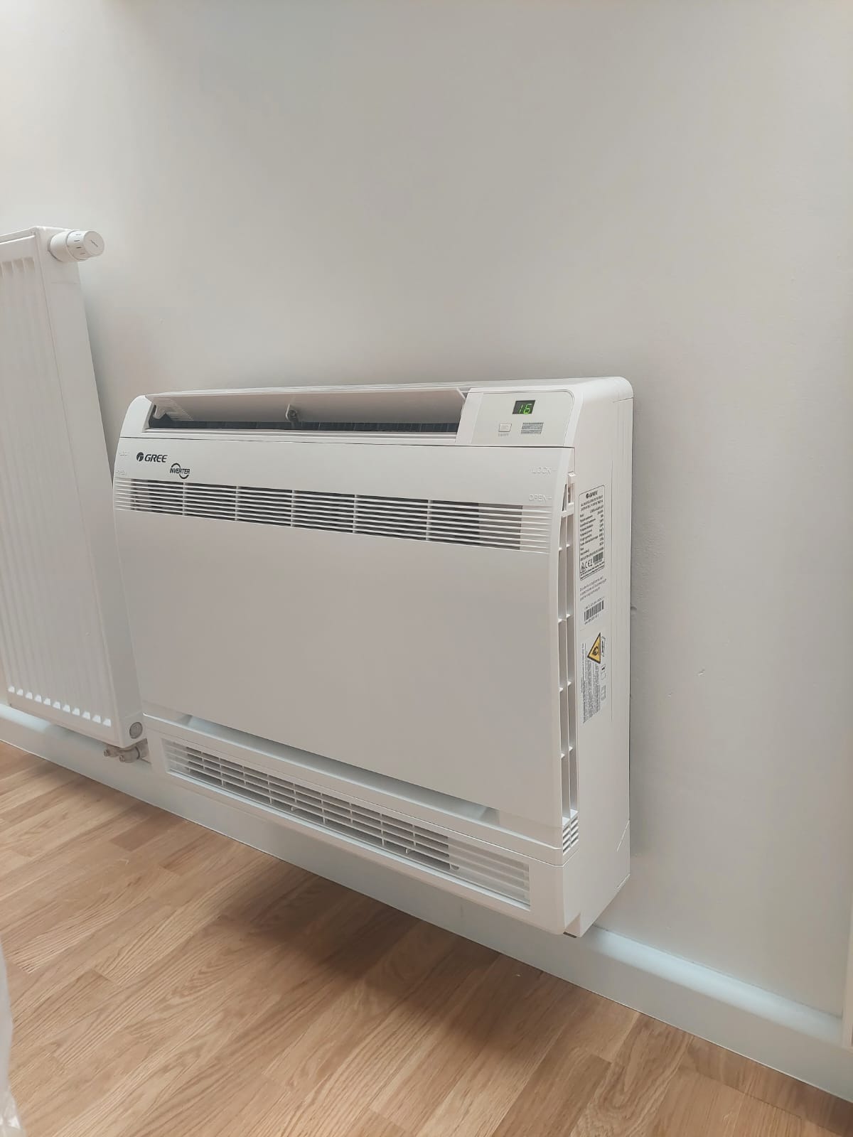 Biała klimatyzacja podłogowa marki Gree zamontowana przy ścianie obok grzejnika, widoczny wyświetlacz z temperaturą.