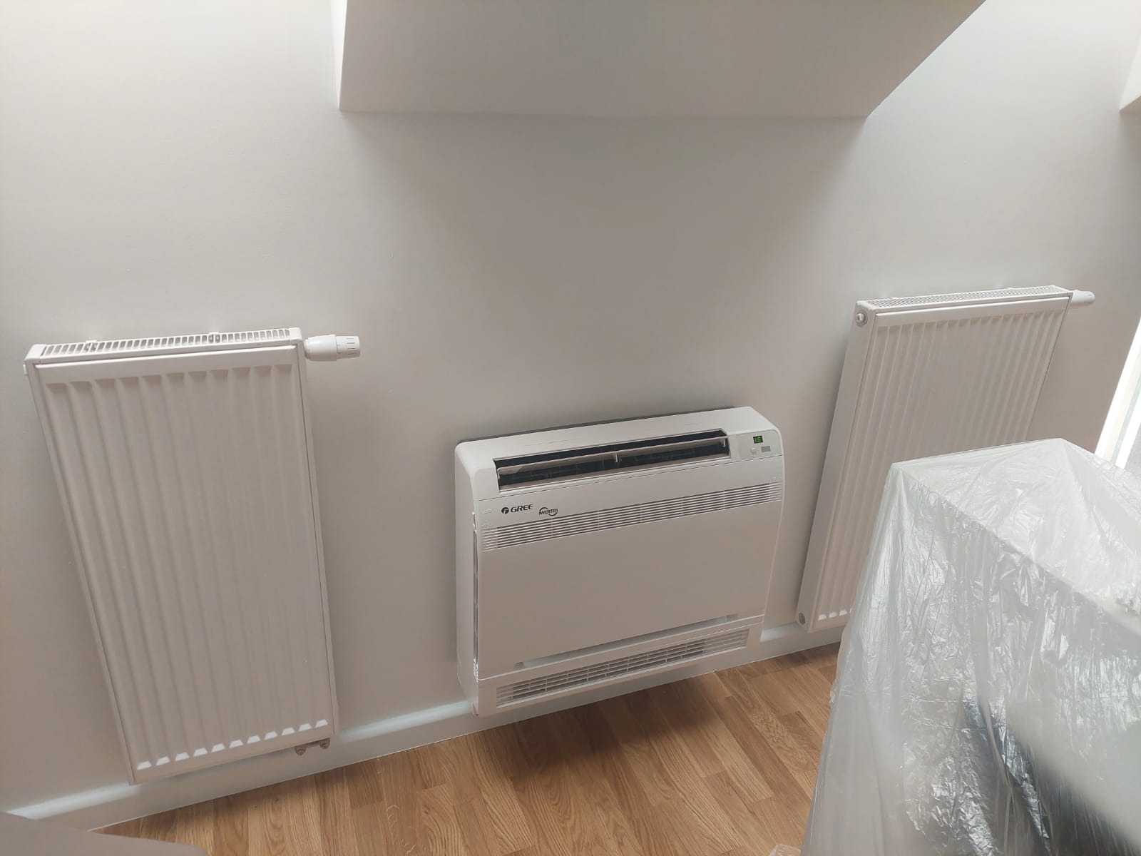 Biały klimatyzator podłogowy marki Gree zamontowany na ścianie w jasnym pomieszczeniu z dwoma grzejnikami panelowymi, pod skosem dachu.