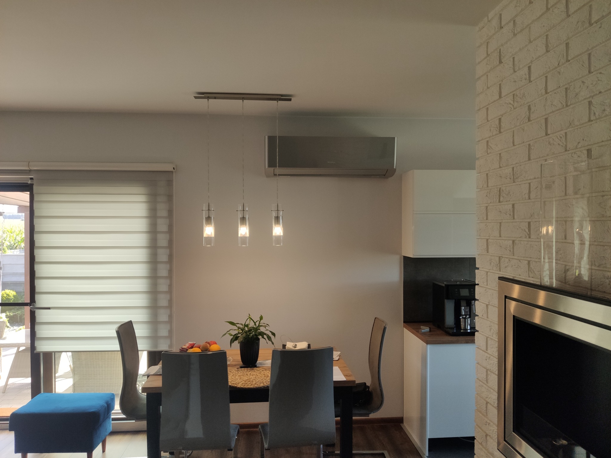 Wnętrze salonu z zainstalowanym klimatyzatorem ściennym marki Samsung, widocznym nad stołem jadalnym z wiszącymi lampami i rośliną w wazonie.