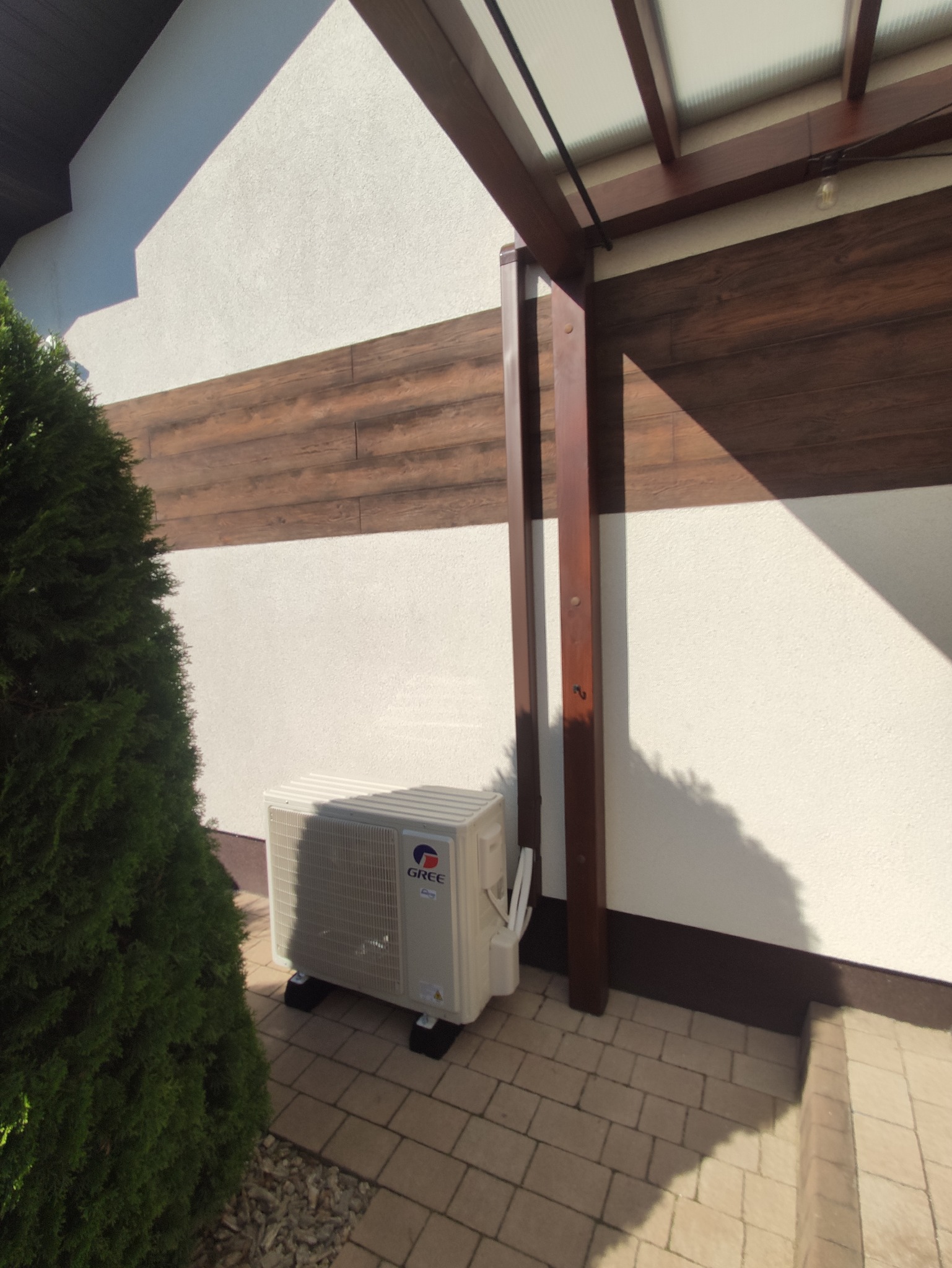 Zewnętrzna jednostka klimatyzacji marki Gree, umieszczona na kostce brukowej przy ścianie budynku z drewnianą elewacją i elementami pergoli.