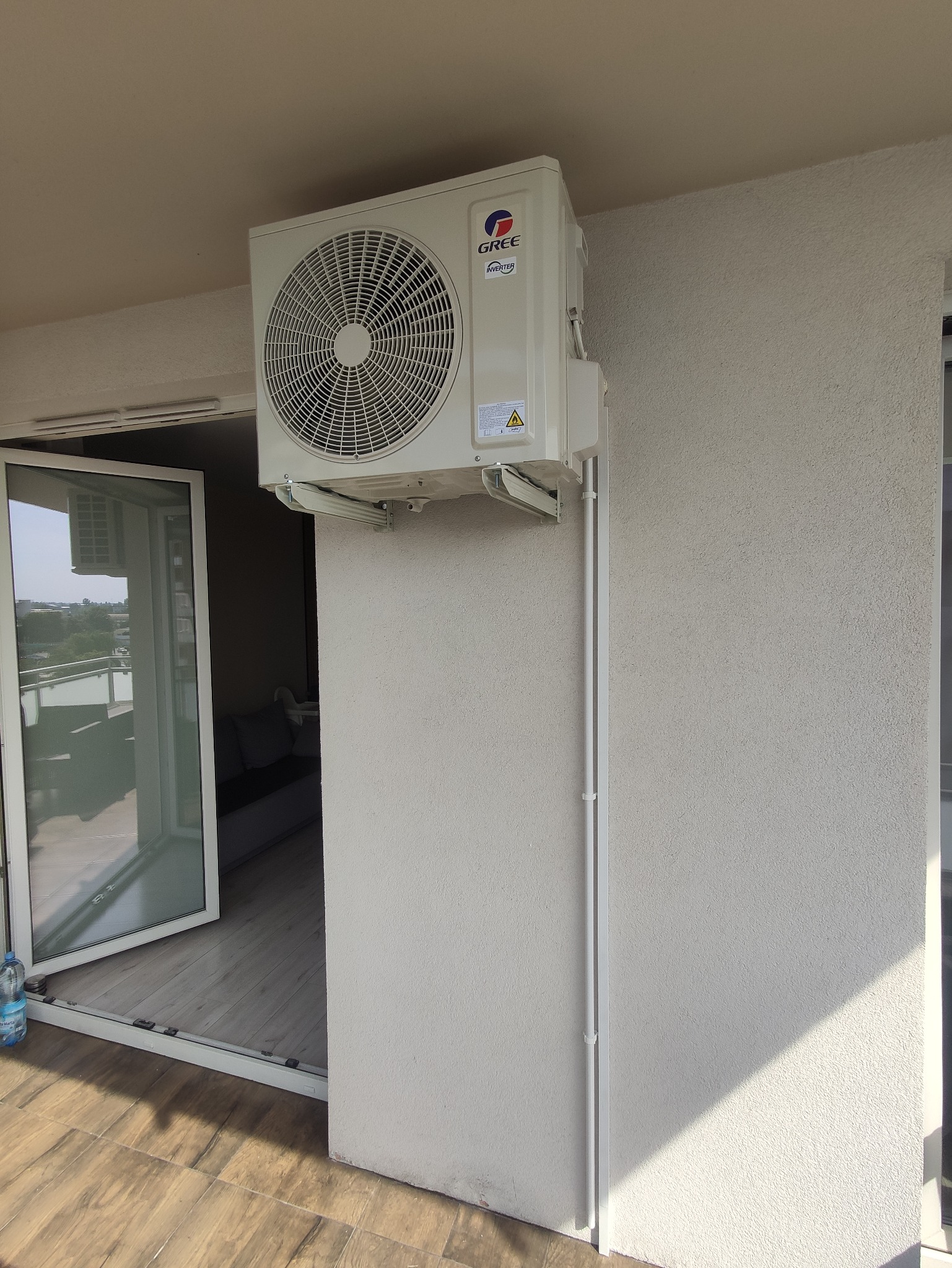 Zewnętrzna jednostka klimatyzacji Gree zamontowana na balkonie, widoczne białe rurki instalacyjne prowadzące wzdłuż ściany, otwarte drzwi balkonowe z widokiem na miasto.