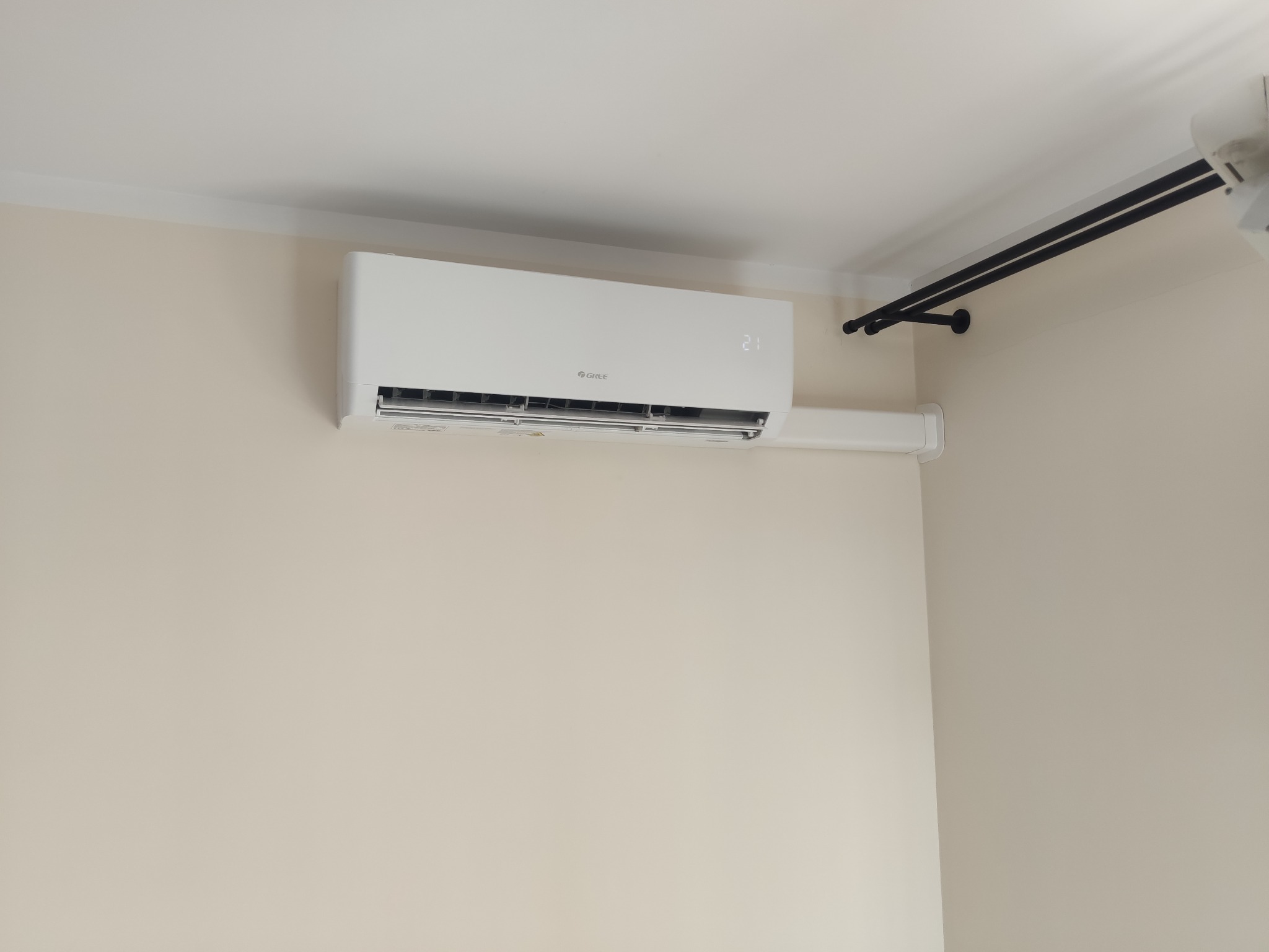 Biały klimatyzator marki Gree z wyświetlaną temperaturą 21 stopni Celsjusza, zamontowany na jasnej ścianie w pomieszczeniu z listwą przysufitową i czarnym karniszem.
