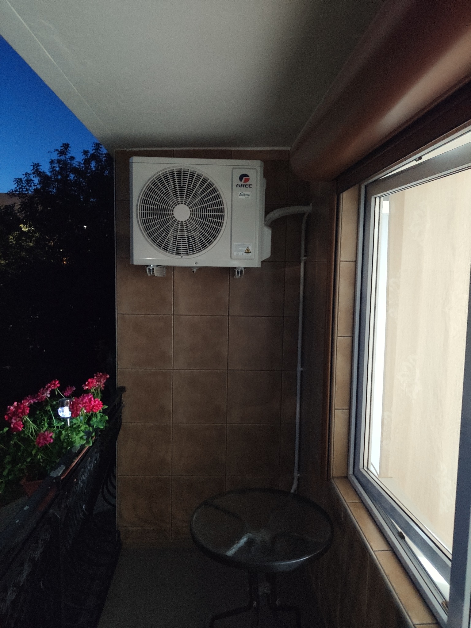 Biała jednostka klimatyzacji marki Gree zamontowana na brązowej ścianie z płytek na balkonie, widoczne kwiaty w doniczce i otwarte okno.