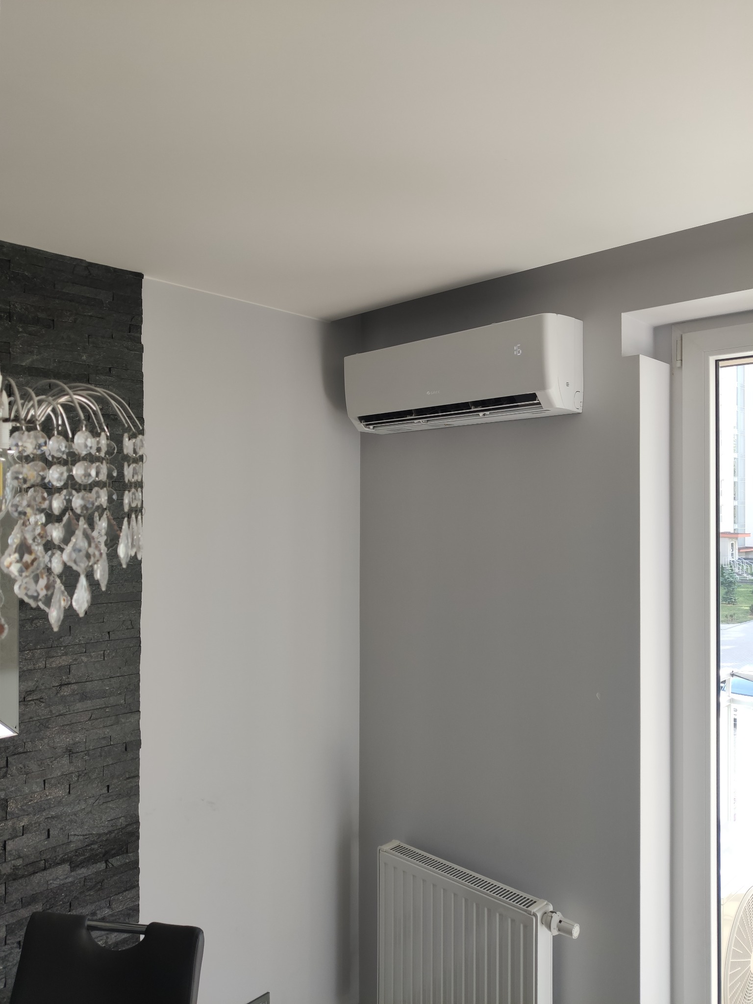 Biały klimatyzator zamontowany na szarej ścianie w nowoczesnym wnętrzu, widoczny grzejnik pod oknem i fragment dekoracyjnej ściany z czarnego kamienia oraz kryształowy żyrandol.