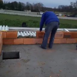 Murarz w niebieskiej bluzie układa cegły na betonowej konstrukcji, w tle widoczny krajobraz z zielenią i szarym niebem.