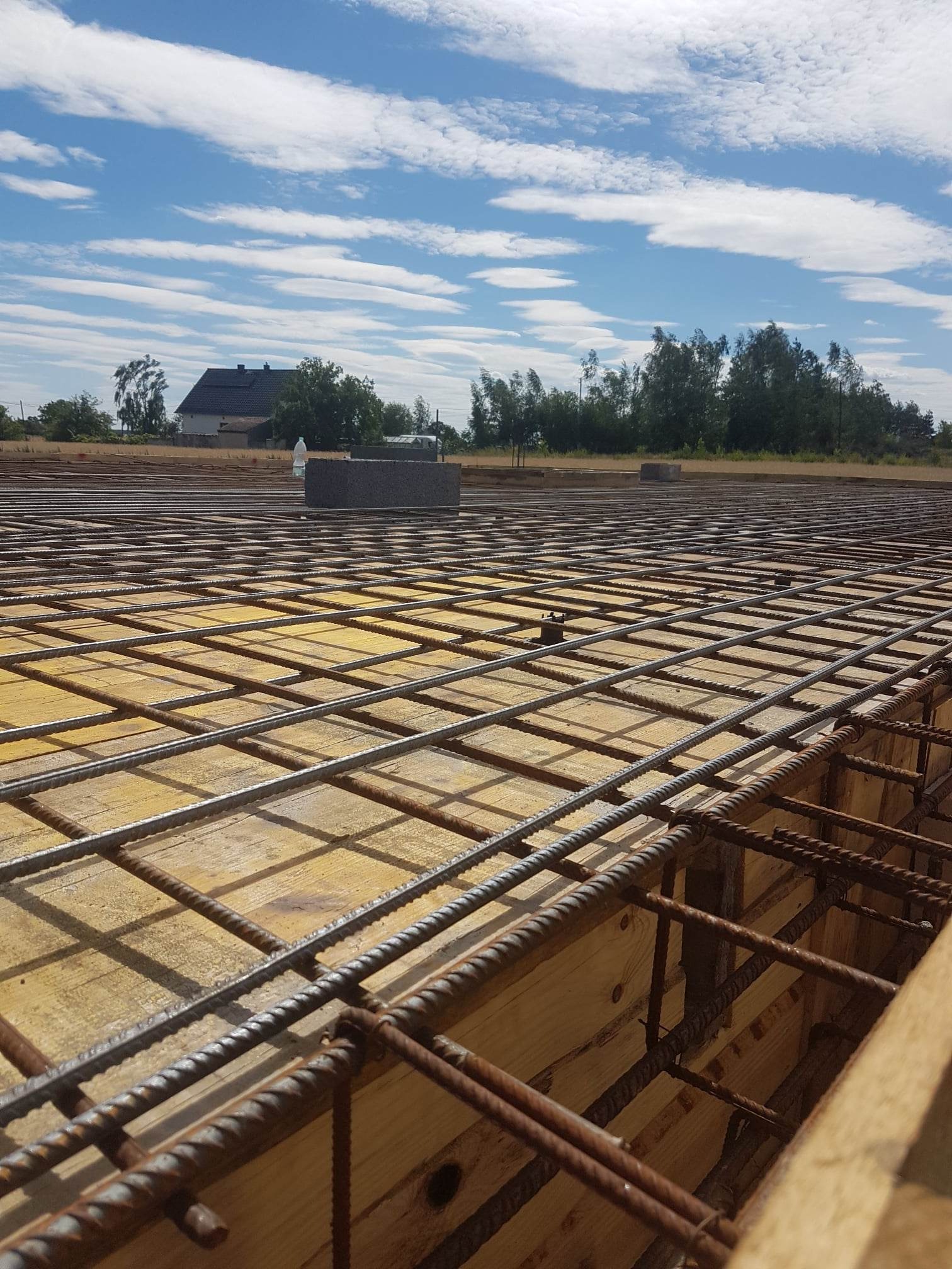 Zbrojenie fundamentu z widoczną siatką stalową na drewnianym szalunku, w tle budynek mieszkalny i błękitne niebo z chmurami.