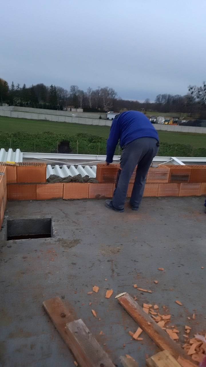 Murarz w niebieskiej bluzie układa cegły na betonowej konstrukcji, w tle widoczny krajobraz z zielenią i szarym niebem.