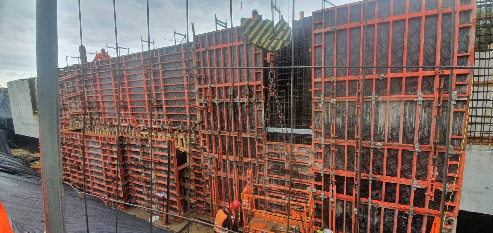 Montaż szalunku zbrojeniowego na budowie, widoczne elementy konstrukcyjne i pracownicy w odblaskowych kamizelkach, dźwig z zawieszoną siatką zbrojeniową.