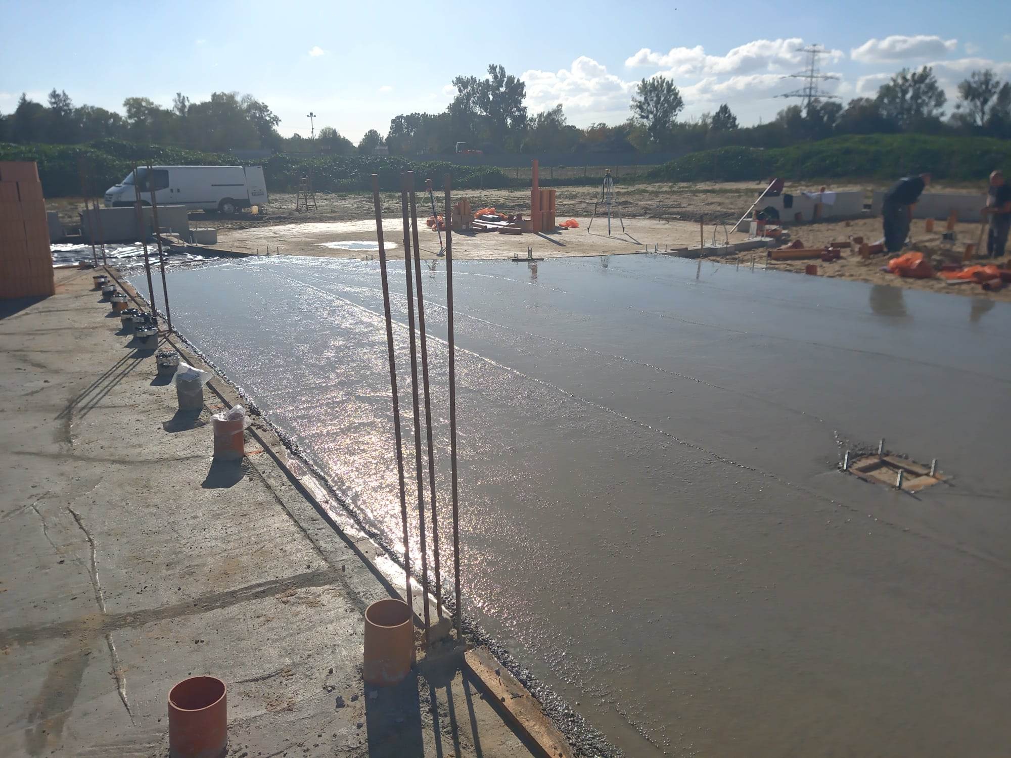 Świeżo wylany beton na budowie, z widocznymi prętami zbrojeniowymi i elementami szalunku. W tle biały bus i pracownicy budowlani.