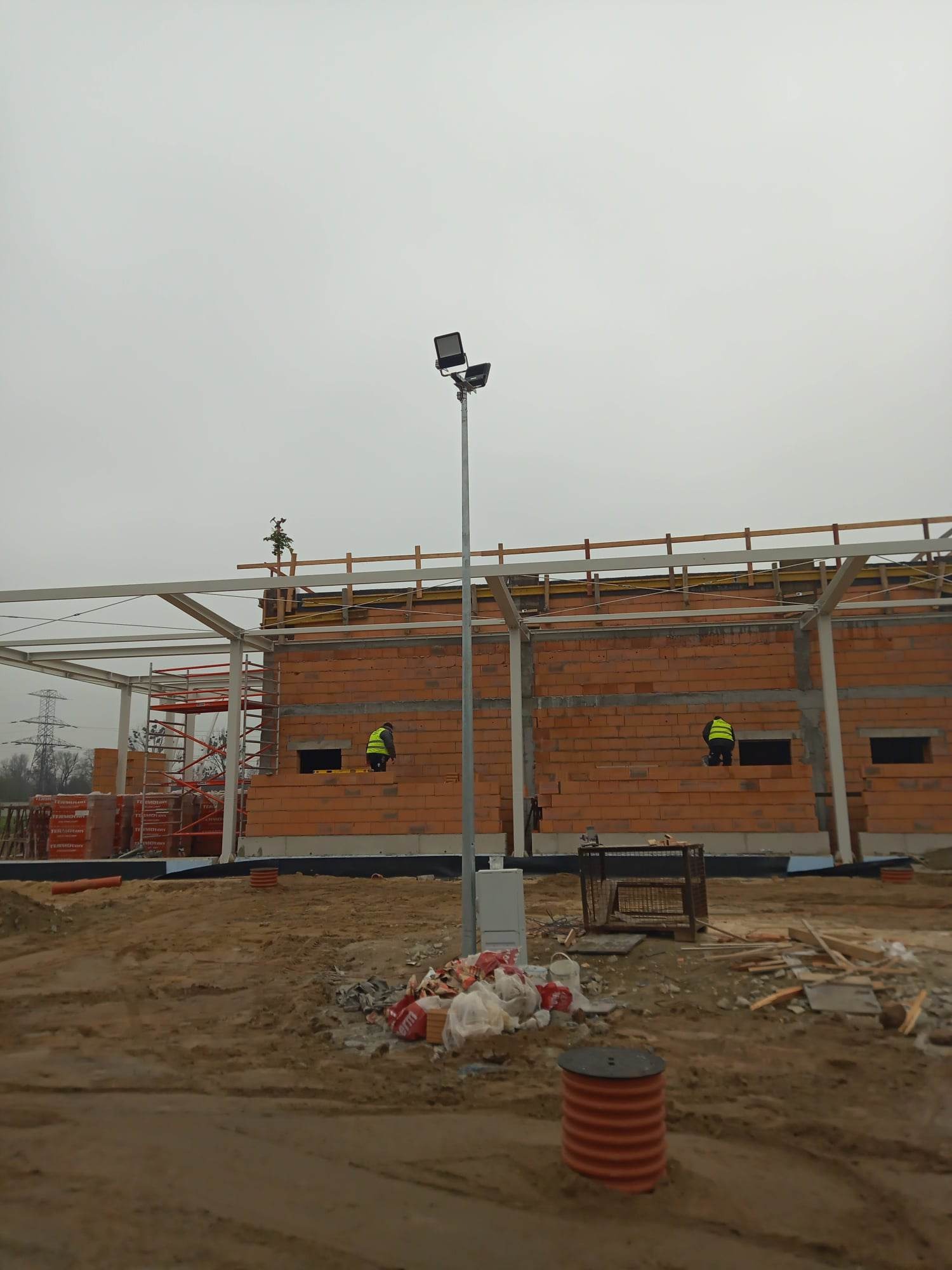 Budowa ściany z czerwonej cegły silikatowej na placu budowy, widoczni dwaj pracownicy w żółtych kamizelkach odblaskowych, jeden na rusztowaniu, drugi przy ścianie. W tle słup z lampami i elementy...