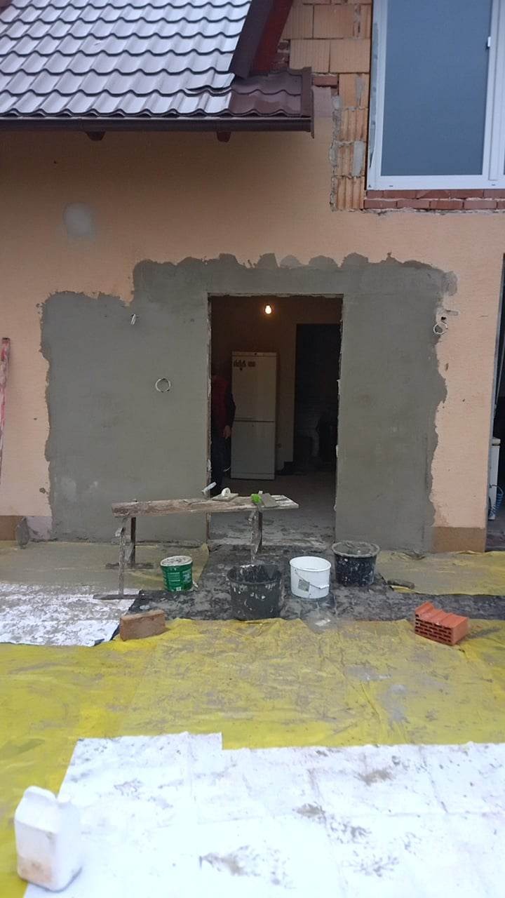 Prace murarskie przy otworze drzwiowym w ścianie zewnętrznej domu jednorodzinnego, widoczne wiaderka z zaprawą, podłoże zabezpieczone folią, w tle fragment elewacji i okno.