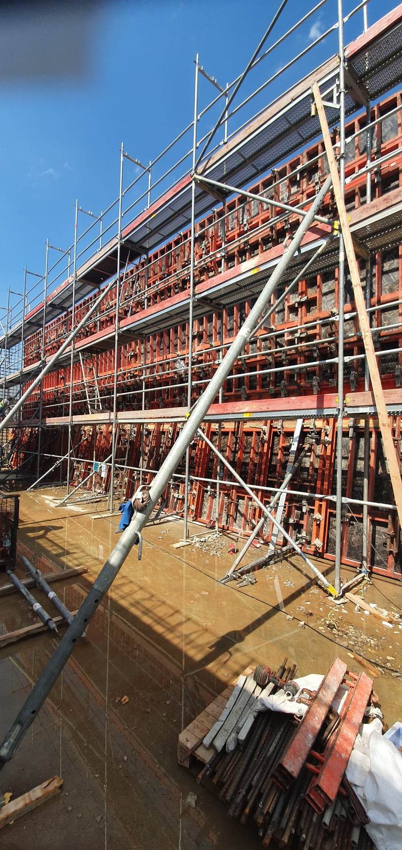 Konstrukcja szalunkowa z rusztowaniami na mokrym placu budowy, przygotowana do wylewania betonu, widok na niebieskie niebo.