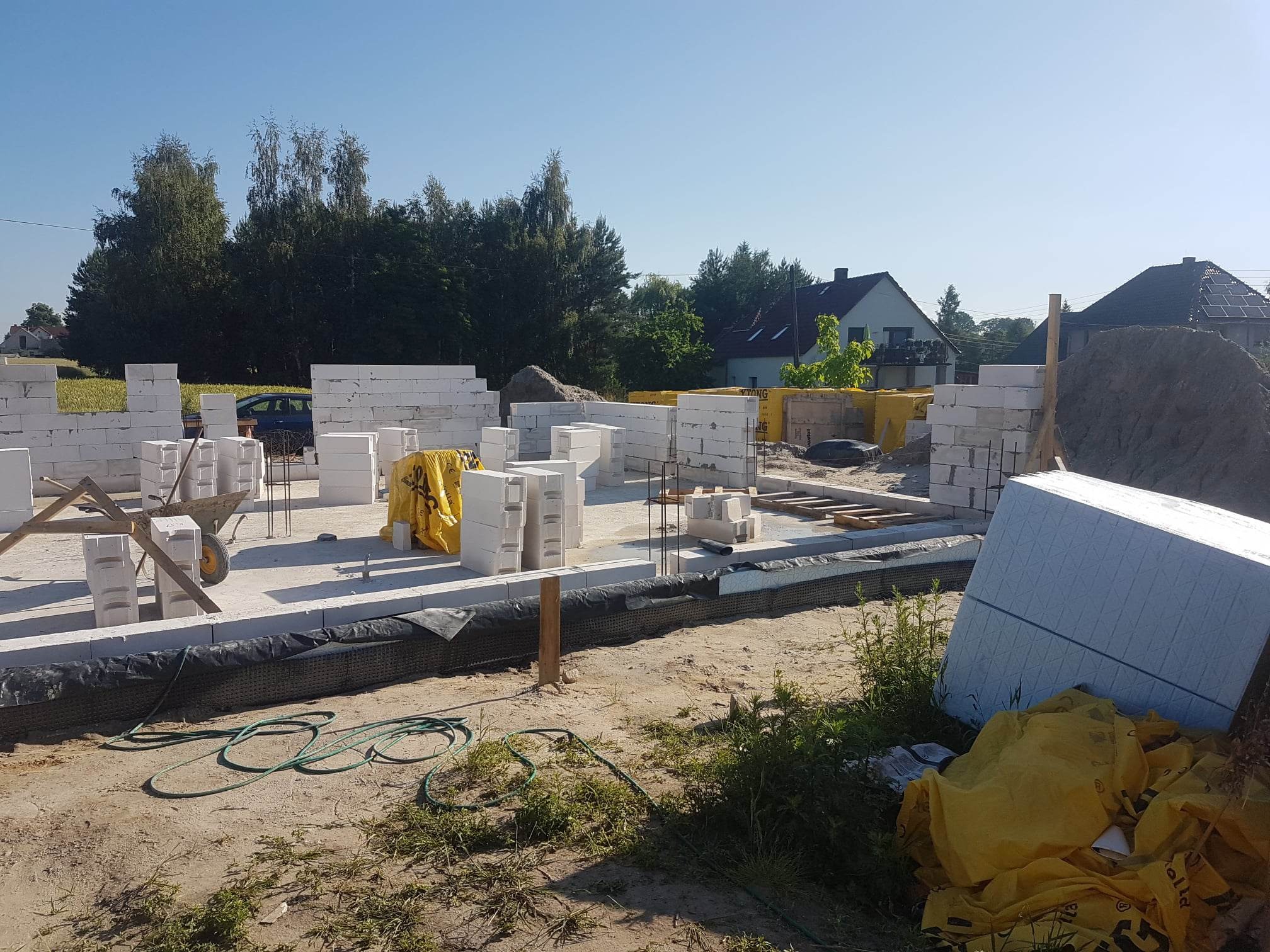 Wznoszenie ścian parterowego domu z bloczków białych, widoczne przygotowane fundamenty, zbrojenie, taczka i materiały budowlane na działce z zielenią w tle.