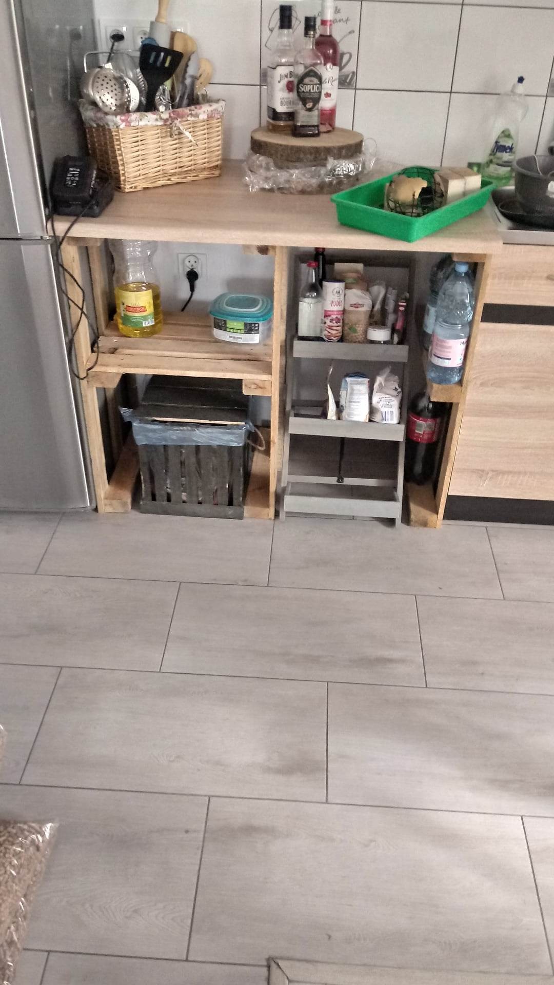 Drewniany regał kuchenny z półkami, wykonany z palet, stojący na szarej podłodze z płytek, z widocznymi produktami spożywczymi i akcesoriami kuchennymi.