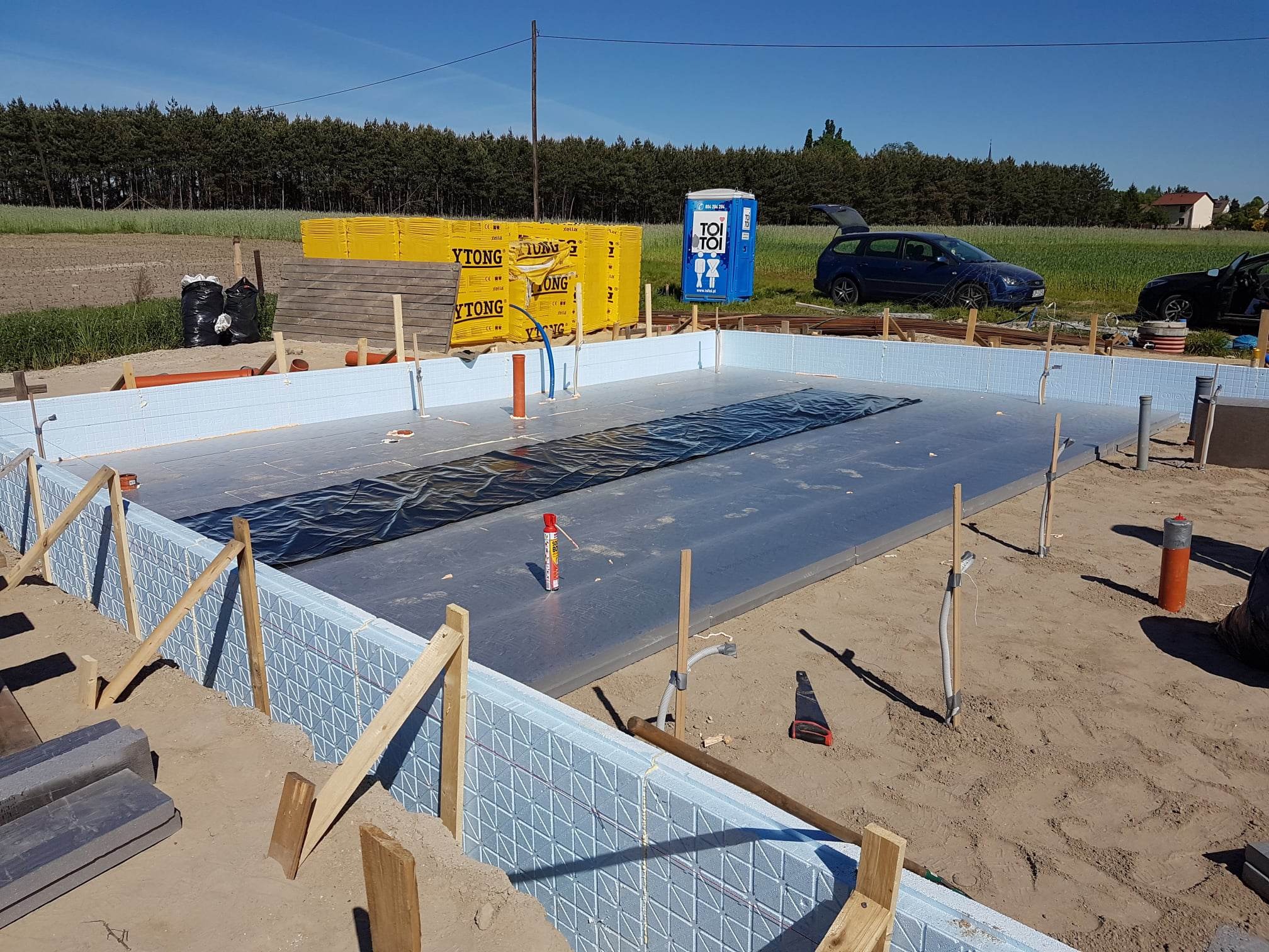 Przygotowanie fundamentów pod budowę domu z wykorzystaniem szalunków z izolacją termiczną, widoczne materiały YTONG, folia izolacyjna, przenośna toaleta TOI TOI i samochody w tle.