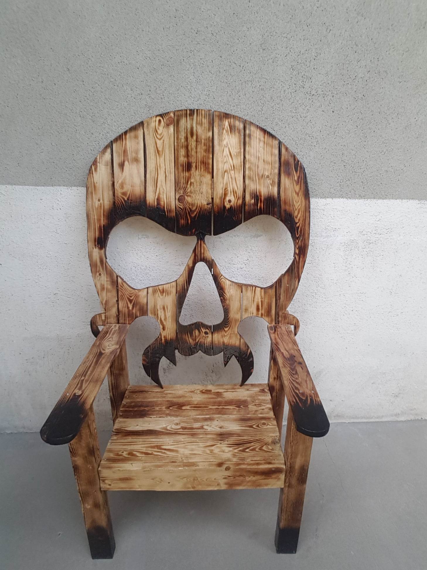 Krzesło wykonane z opalanych desek, z oparciem w kształcie czaszki, stojące na tle szarej ściany.