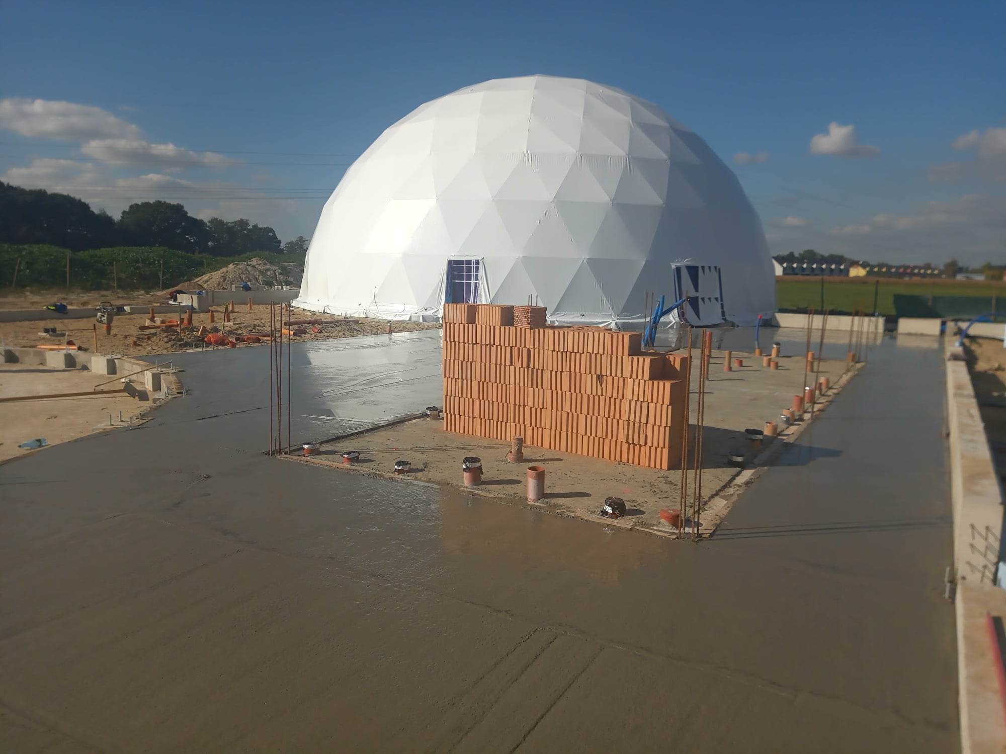 Budowa nietypowego budynku o konstrukcji kopułowej z białego materiału, widoczny wylany betonowy fundament i paleta czerwonej cegły, niebieskie niebo w tle.